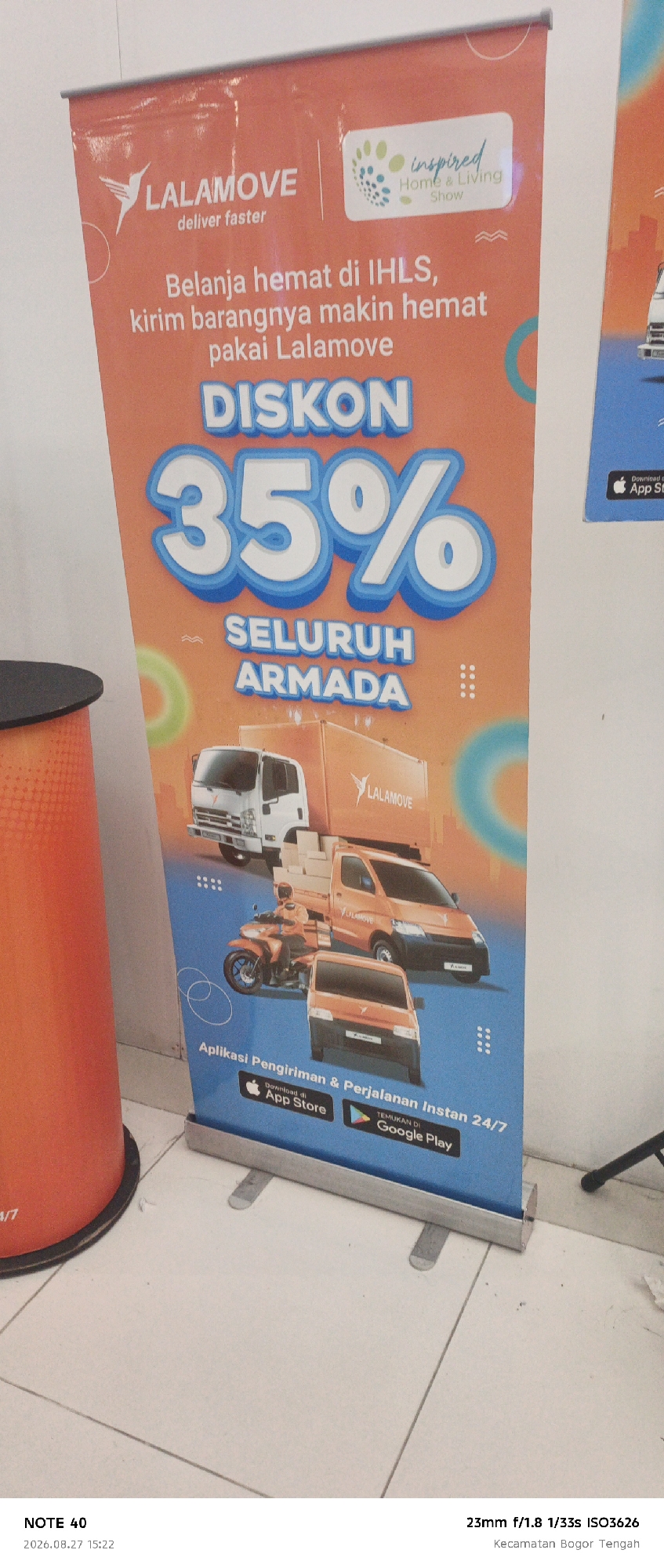The image shows a banner with a promotional advertisement for LALAMOVE, a delivery service. The banner features graphics of delivery vehicles, including a truck, a van, and a motorcycle with a rider. It highlights a "DISKON 35%" (35% Discount) for "SELURUH ARMADA" (Entire Fleet). There's also text in Indonesian that translates to "Shop affordably at IHLS, ship your goods even cheaper with Lalamove" and mentions "Aplikasi Pengiriman & Perjalanan Instan 24/7" (Instant Delivery & Travel App 24/7) with icons for App Store and Google Play. The banner is positioned in what appears to be an indoor exhibition or event space, possibly a mall or convention center, with a tiled floor. In the bottom left corner, text indicates "NOTE 40 2026.08.27 15:22" and "23mm f/1.8 1/33s ISO3626 Kecamatan Bogor Tengah," suggesting the photograph was taken in Bogor Tengah, Bogor, Indonesia on August 27, 2026, at 3:22 PM.