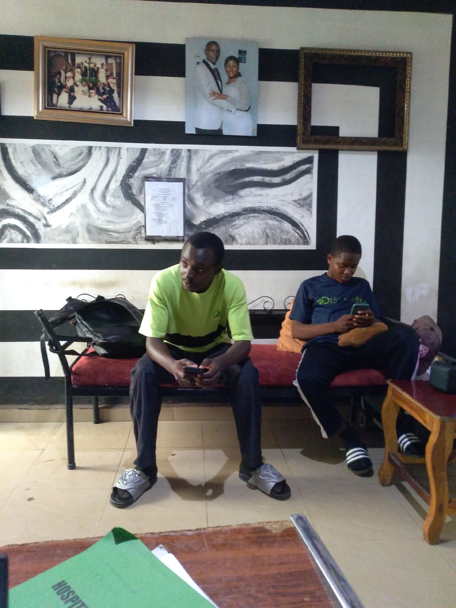 The image depicts an indoor scene with two young Black males seated on a bench. The male on the left wears a lime green t-shirt with black stripes and dark pants, holding a smartphone. The male on the right wears a dark blue t-shirt with white lettering and dark track pants, also holding a smartphone. Both are wearing casual footwear. Behind them, a black and white striped wall serves as a backdrop, adorned with framed pictures and artwork, including what appears to be a wedding photograph. A dark backpack rests on the bench next to the male on the left. A wooden table with a red top is positioned to the right, holding a small green object with the word "HOSPIT" visible. The floor is tiled. The environment suggests a waiting area or lounge, possibly within a business or public institution in Jos, Nigeria.