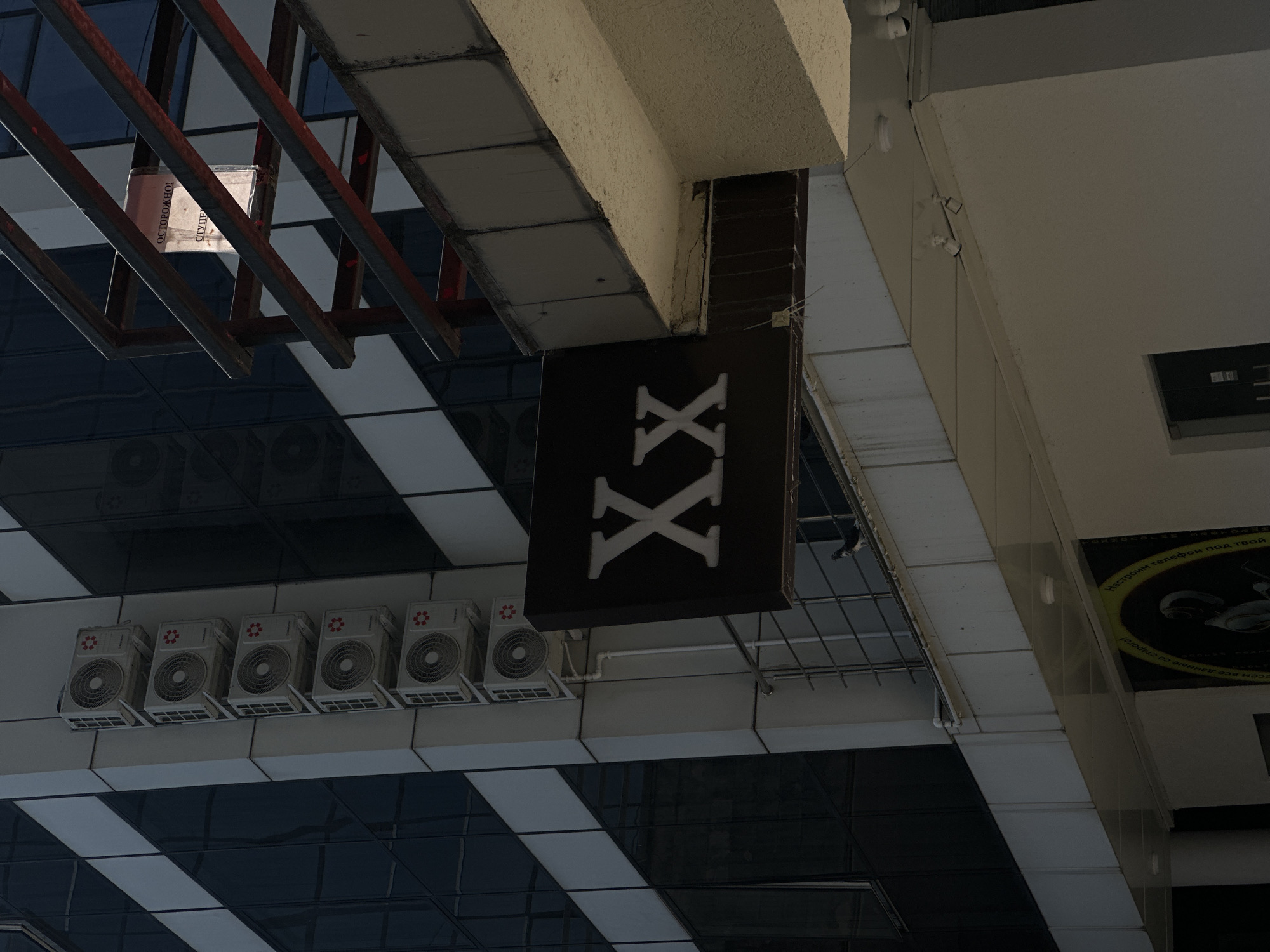 The image shows the exterior of a building in Sochi, Russia. A dark sign with white "Xx" lettering is mounted on the side of the building. Above this sign, there are several white air conditioning units stacked vertically on the building's facade. To the right of the sign, a metal railing structure is visible, with a sign that appears to be warning of danger in Russian. The building has large dark windows. The lower part of the image shows a white awning or overhang, with some recessed lighting fixtures visible. A bird is perched on a railing in the foreground. The overall scene suggests an urban commercial or office building environment.