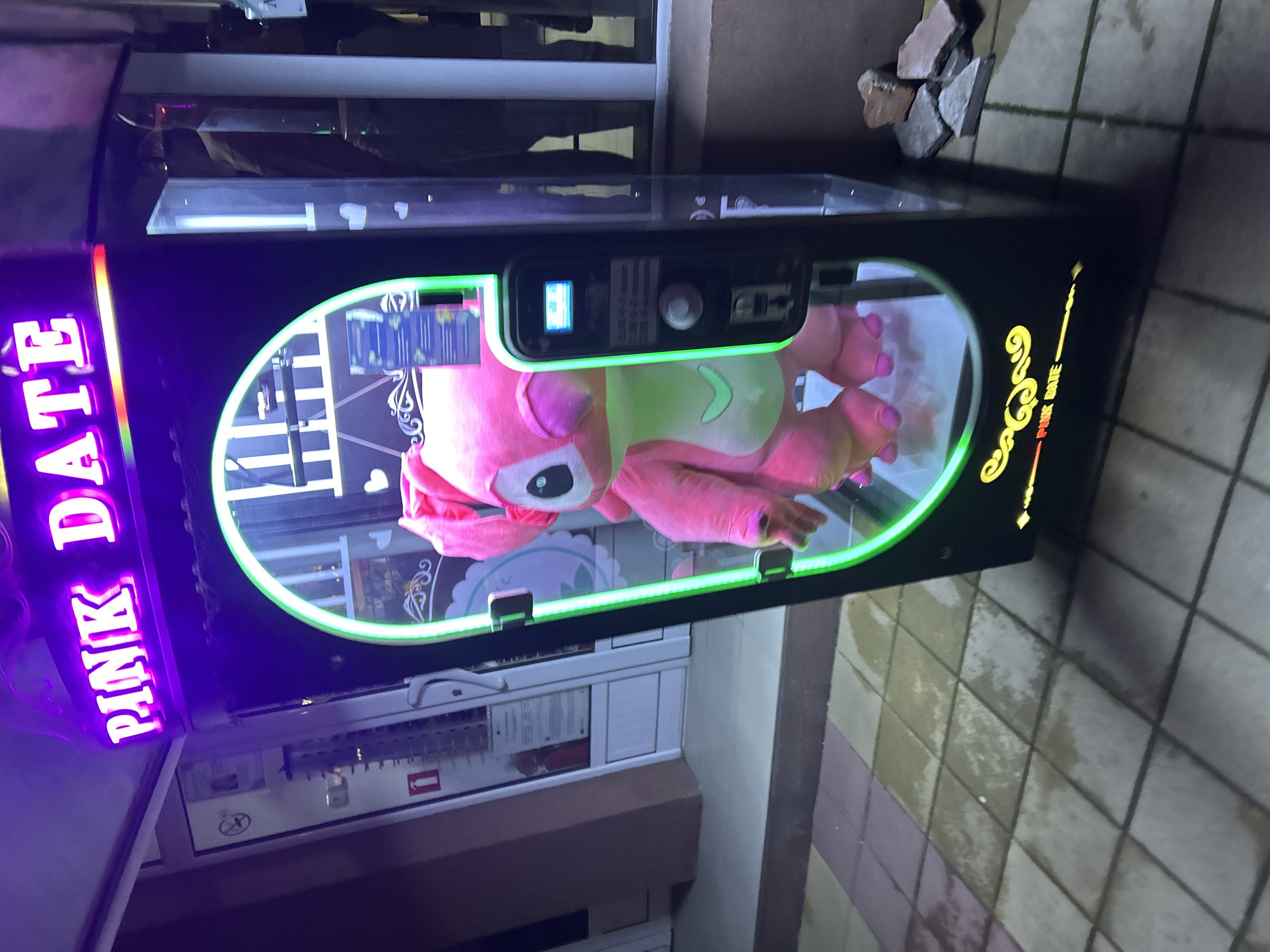 The image depicts a claw machine, labeled "PINK DATE," located in Zlatograd, Bulgaria. The machine houses a pink stuffed animal inside a glass enclosure, surrounded by a green neon light. The machine is positioned on a tiled walkway near a building entrance. The scene appears to be outdoors.