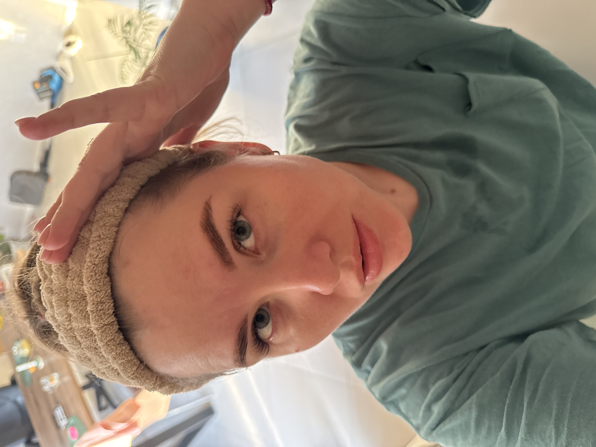 A woman with light skin, light eyes, and dark eyebrows is looking at the camera. She is wearing a beige headband. Her left hand is raised to her forehead. She is wearing a light green sweatshirt. The background is blurry and includes what appears to be a window and some objects on a shelf. There is no location provided to me in Ферма, Russia.