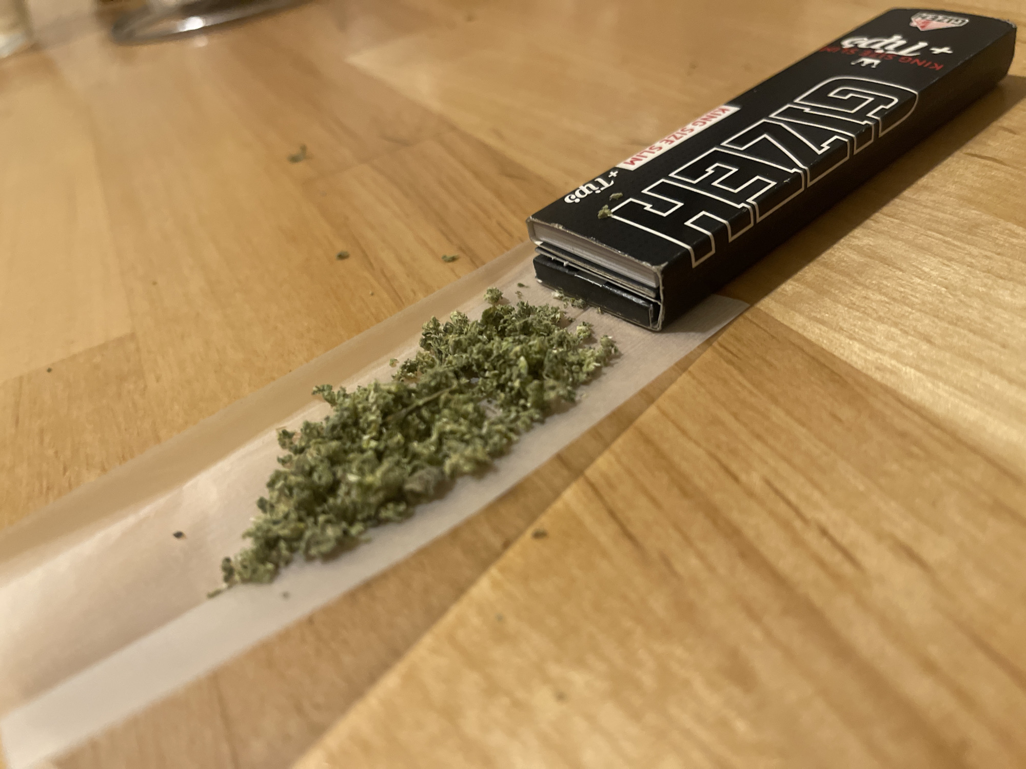 The image captures a close-up, eye-level view of items for rolling a cigarette or joint, set against a warm-toned wooden surface, likely a table or floor. The central focus is on a white, translucent rolling paper, upon which a pile of finely ground green material, consistent with cannabis, is distributed in preparation for rolling.
To the right of the rolling paper lies a black packet of rolling papers and tips. The text "GIZEH" is prominently displayed in large white letters on the packet, along with "KING SIZE SLIM" and "+ Tips" in smaller font. A small logo, possibly a crown, is also visible on the packet. Several tiny specks of the green material are scattered on the wooden surface around the main pile, suggesting some spillage during preparation. In the upper left background, a blurry metallic object is partially visible, offering a hint of other items in the environment. The lighting suggests an indoor setting, but the time of day is not discernible. The scene implies the activity of preparing to roll and consume the green material.