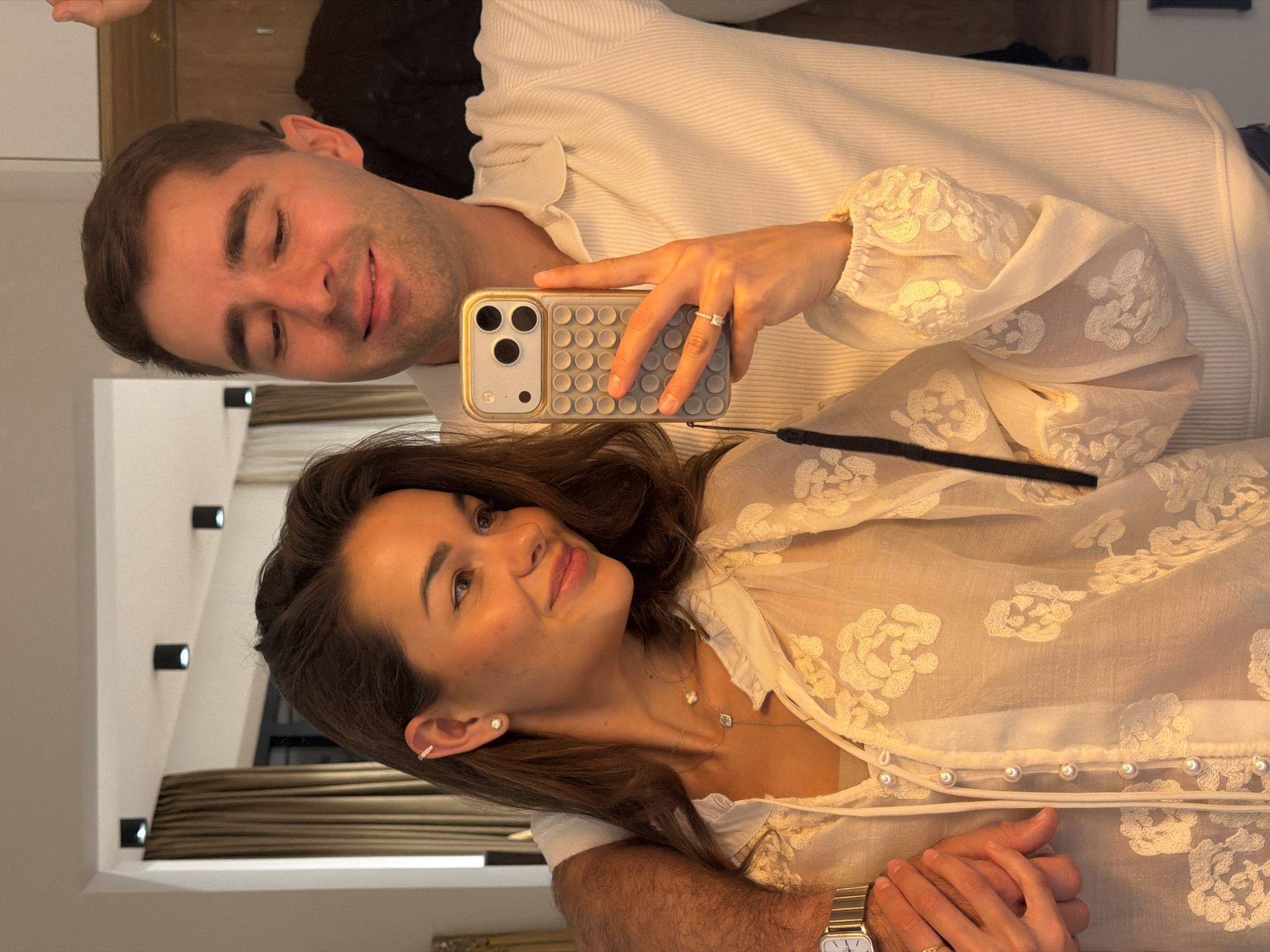 An indoor mirror selfie taken in Tbilisi, Georgia, depicts a man and a woman. The woman, positioned on the right, holds a silver/gold iPhone in a grey textured case with a grid pattern. She has dark hair, wears a sheer, light-colored blouse with white floral embroidery and pearl buttons, and accessorizes with a stud earring, an ear cuff, two delicate necklaces (one with a square pendant, one with a heart), and a ring on her left ring finger. She is smiling, looking towards the man's reflection. The man, standing behind her with his arm around her shoulder (partially visible), wears a light-colored, ribbed polo shirt and smiles downwards. His left wrist displays a rectangular watch with a metal band. Their hands are intertwined in the lower-left corner of the frame. The background, visible through the mirror, reveals an interior space with light-colored walls, three black cylindrical ceiling spotlights, and light brown curtains.