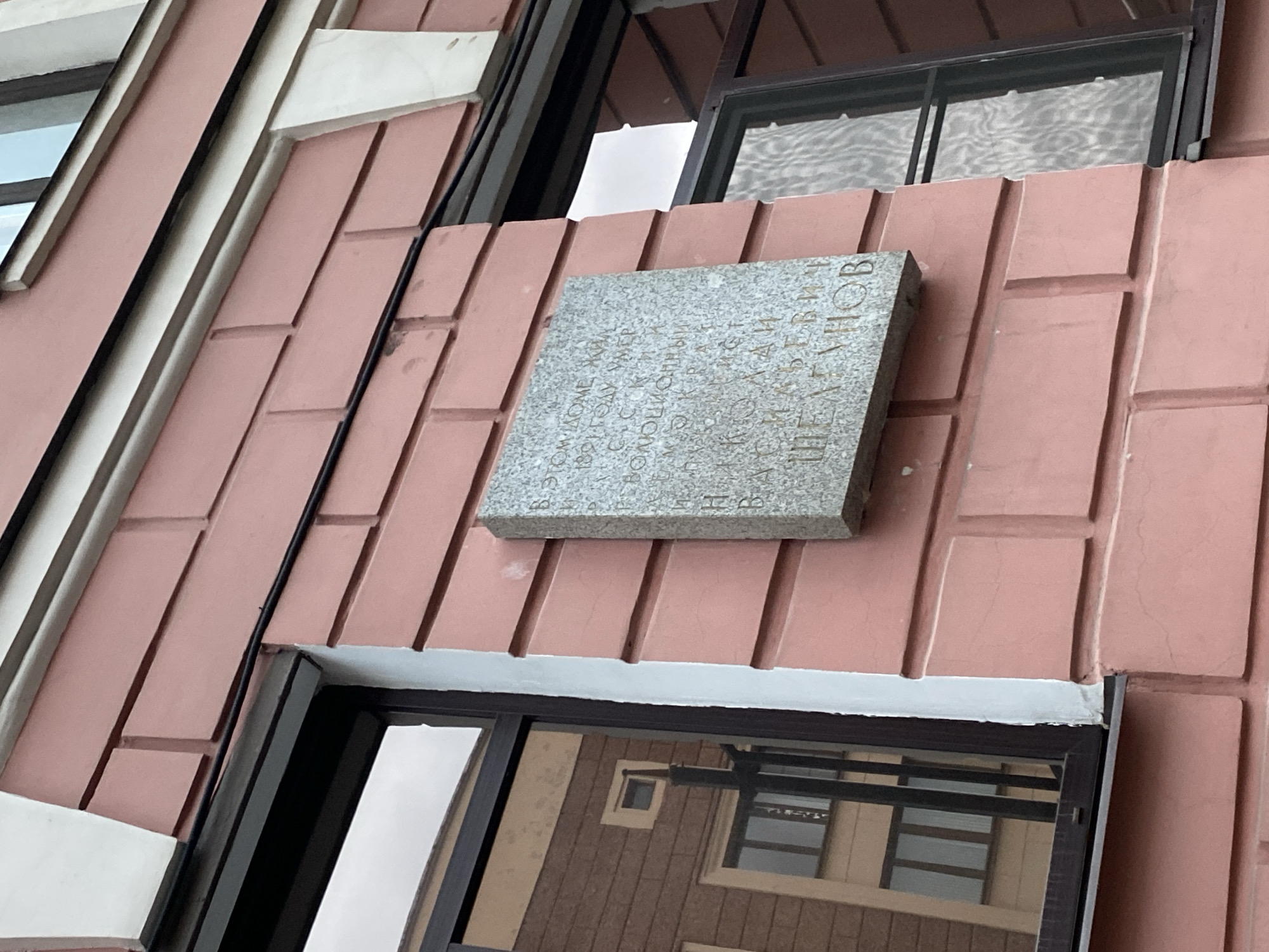The image shows a granite memorial plaque mounted on the exterior wall of a building in Saint Petersburg, Russia. The building facade is clad in a reddish-brown material with a grooved, modular pattern. The plaque is gray with text in Russian. A portion of windows with black frames are visible above and below the plaque, reflecting the surrounding environment. The architecture appears to be modern or post-Soviet.