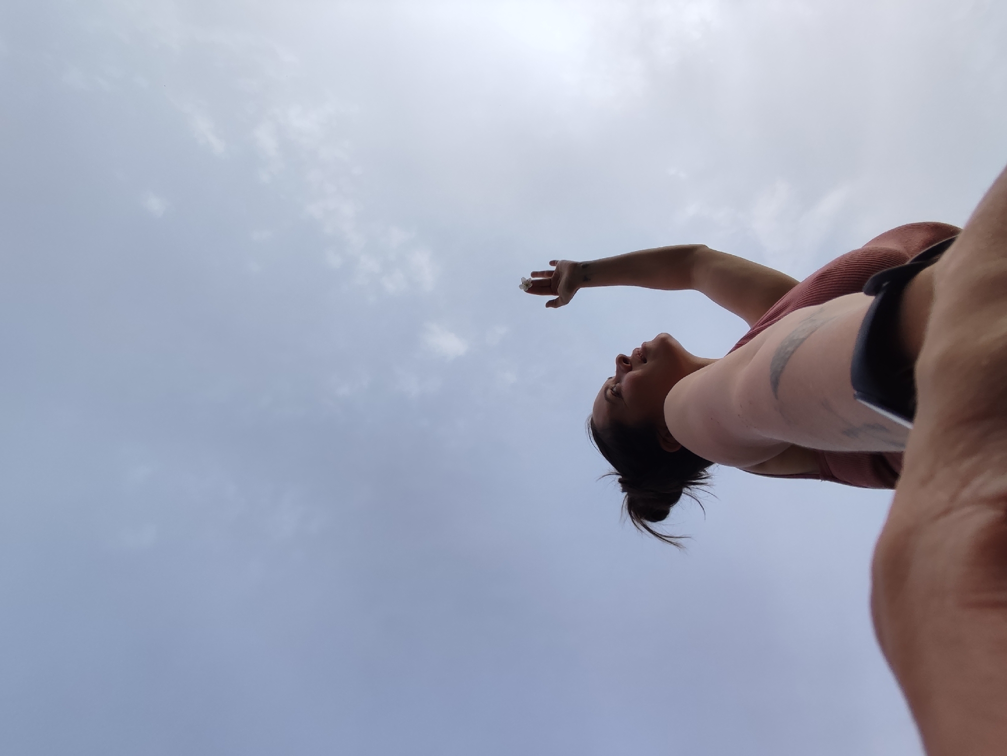 A low-angle shot shows a woman looking up toward the sky, extending her arm and hand. The sky is overcast. The woman has dark hair pulled back in a bun, a pink top, and a dark band visible on her arm. A tattoo is visible on her arm. No information indicates this image was taken in the city of Фоки, Russia.