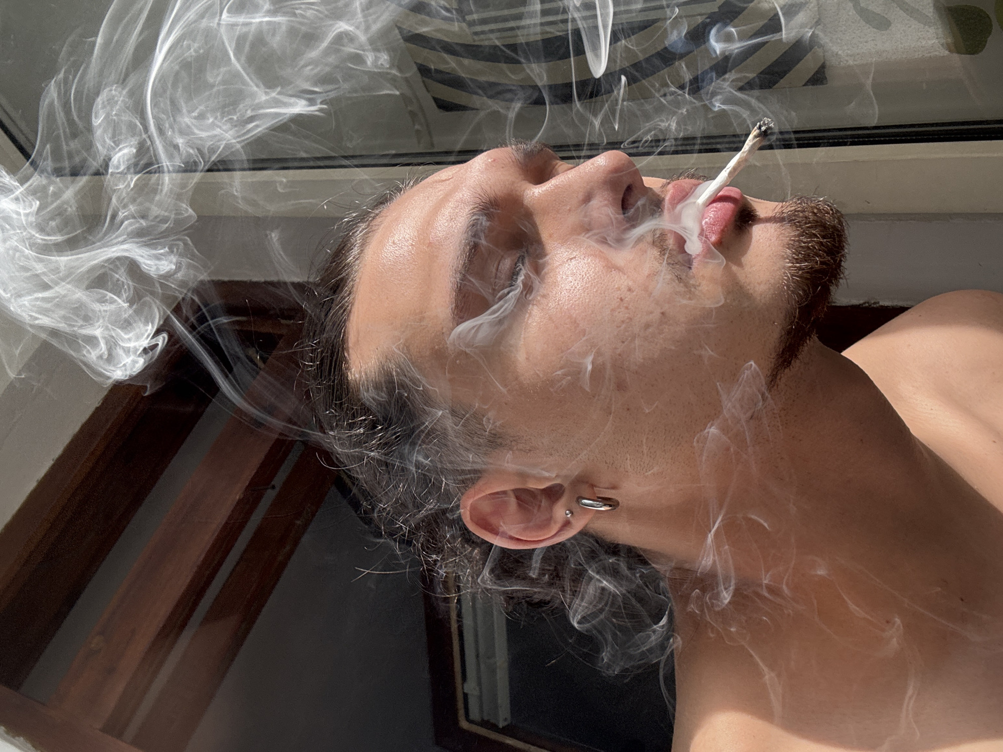 A close-up, profile shot captures a young person with their eyes closed, exhaling smoke from a cigarette held to their lips. The subject has dark, curly hair pulled back, a hoop earring in their left ear, and a light beard and mustache. Smoke billows around their face and across the frame, creating an ethereal effect. The background appears to be a window with a view of what could be a city, with vertical blinds visible through the glass. The lighting suggests it is daytime, possibly late afternoon, given the warm, golden hue and the shadows. The setting does not provide specific visual cues to identify the location as Sofia, Bulgaria; it is a generic urban interior with a view.