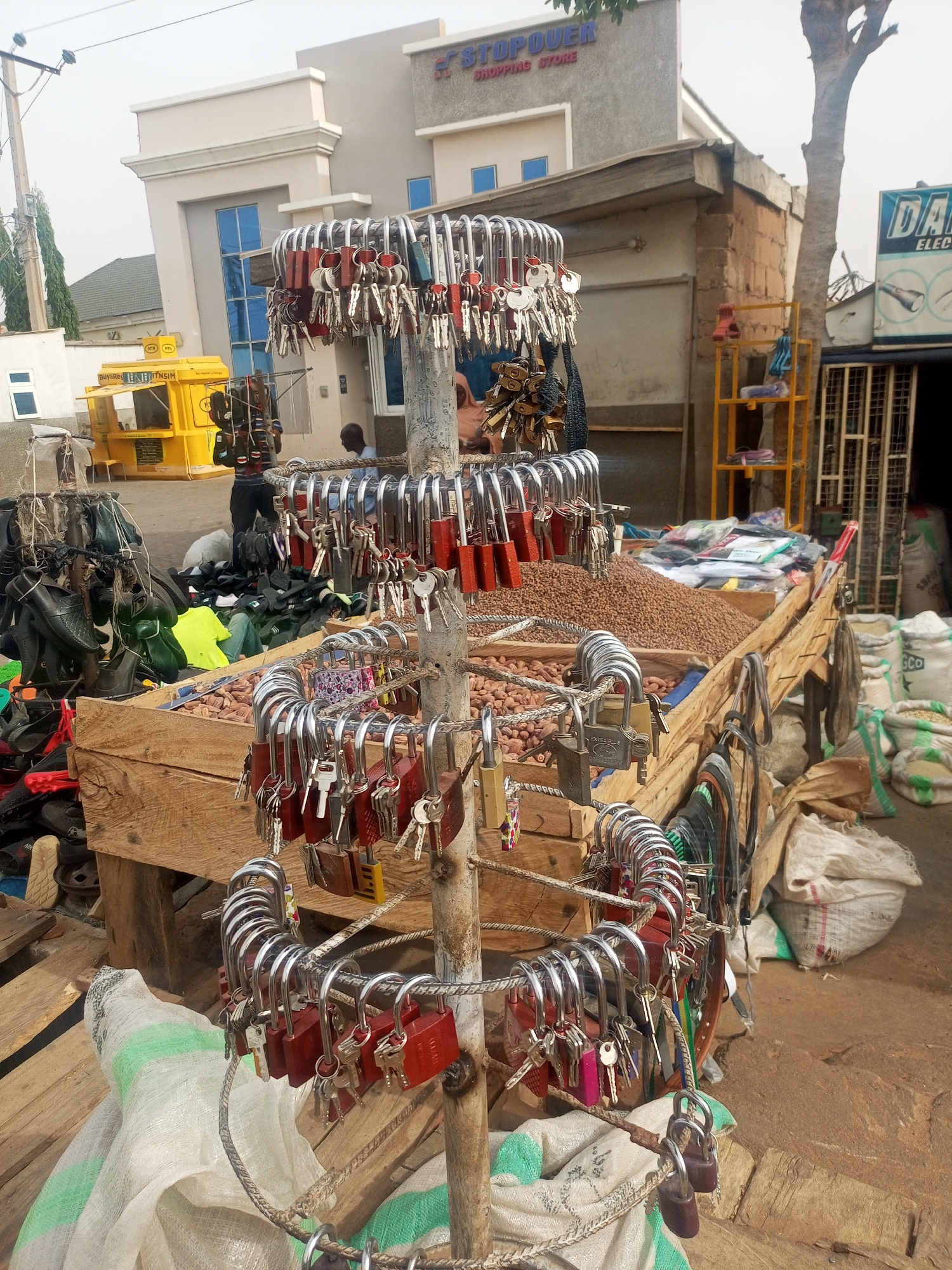 Captured in the Nigerian city of Tempure, the image portrays a bustling street-side market scene. A stand constructed from wood serves as the focal point, displaying a large mound of brown peanuts or legumes.
Suspended around a vertical metal pole are numerous padlocks with their accompanying keys, seemingly offered for sale. To the left, there is an assortment of footwear, including shoes and sandals. Sacks and bags are scattered around the wooden structure.
In the background are several buildings and structures: one with "STOPOUER SHOPPING STORE" signage, a yellow kiosk, and other commercial establishments. A few individuals are visible in the midground and background, and the environment appears dusty.