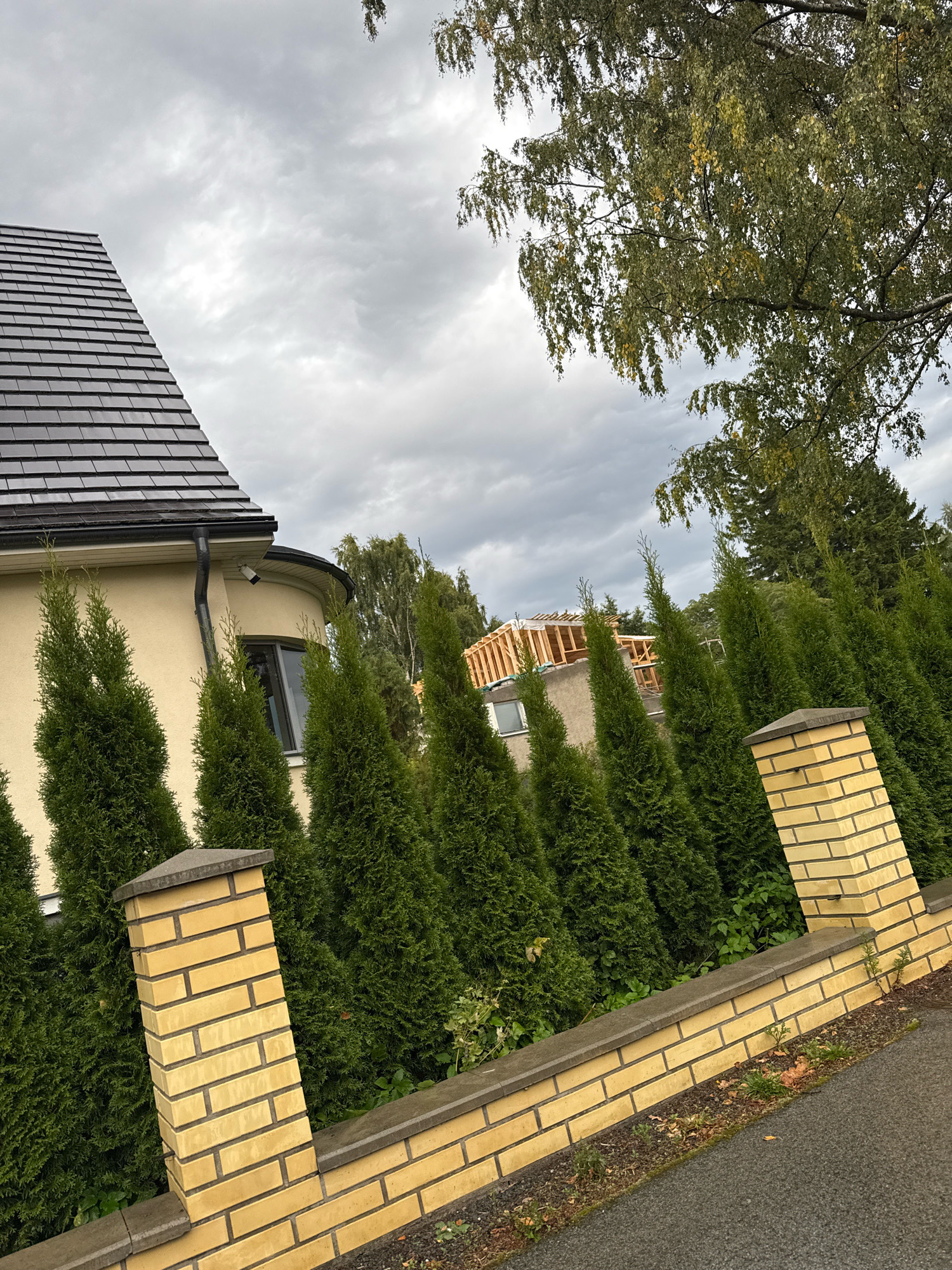 The image depicts a residential property with a house, a garden fence, and landscaping under an overcast sky. A portion of a beige house with a dark grey shingled roof is visible on the left. A black downspout runs along the side of the house, and a security camera is mounted near the eaves. A curved section of the house's exterior wall is also visible, with a dark-framed window.
In front of the house, a manicured row of dark green, conical evergreen trees forms a hedge. A low, yellow brick fence with grey stone capping runs along the foreground, separating the property from a grey asphalt pathway. The fence has brick pillars topped with grey stone caps. Behind the hedge, a partially constructed wooden structure, likely an extension or a new building, is visible. Trees with green and yellowing leaves, suggesting autumn, are present in the background. The overall scene suggests a suburban or residential area. The image appears to be taken in Tallinn, Estonia, based on common architectural styles and landscaping found in the region.