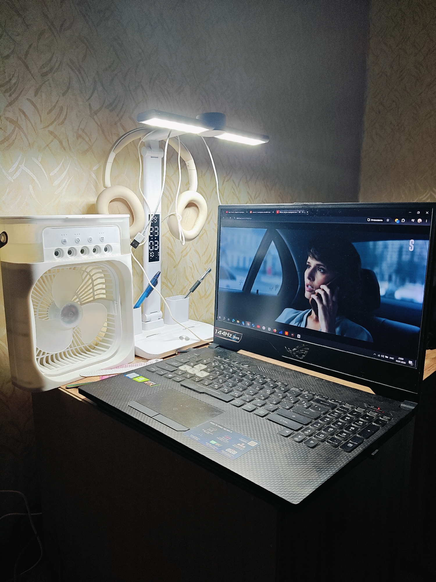 A bedside desk arrangement is captured in a dimly lit room, with a patterned wallpaper in the background. On the left, a white portable fan with multiple settings is positioned. To its right, a white desk lamp with a flexible arm illuminates the area. Two pairs of beige headphones hang from the lamp. Next to the lamp, a white pen holder contains a blue pen and a grey pen. A laptop sits open in the center, displaying a scene from a movie or TV show featuring a woman on a phone call inside a car. The laptop's keyboard is visible, showing Russian characters. The surface of the desk is made of wood with a visible grain. The scene appears to be in an indoor setting, possibly a bedroom or living space. The date stamp on the laptop screen indicates "10.07.2026", suggesting a future date. There is no explicit indication of the city of Chaikovsky, Russia, within the image itself.