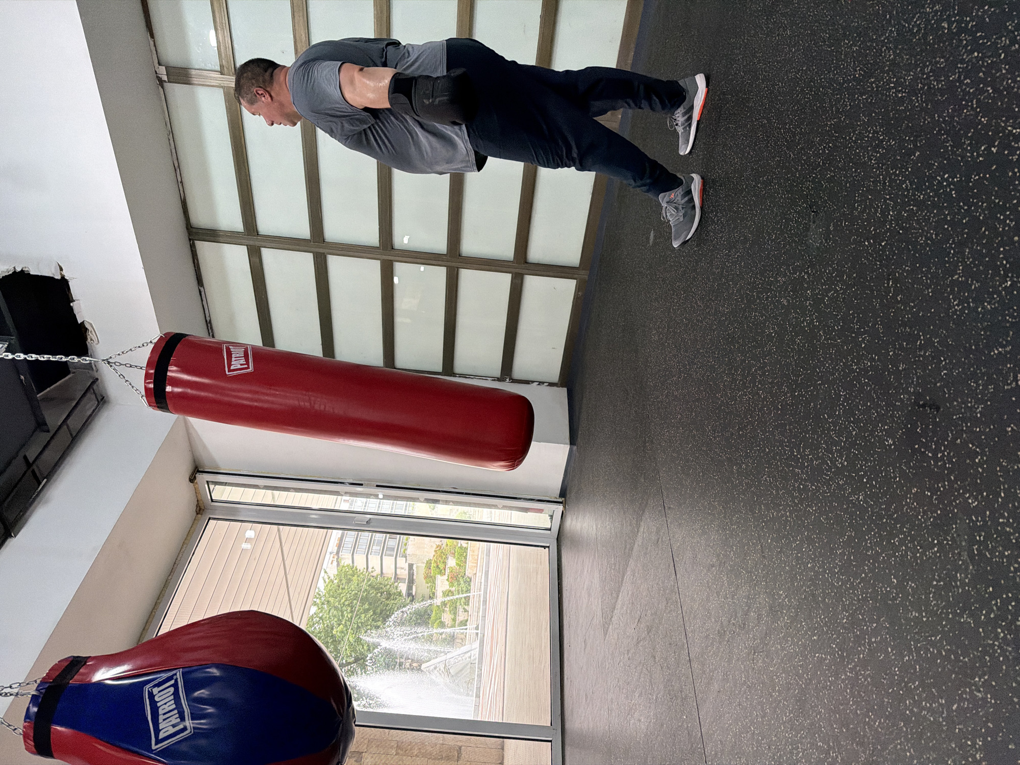 The image shows a man performing a plank exercise in an indoor gym, located in Blagoevgrad, Bulgaria. The man, appearing to be in his 30s or 40s, is wearing a grey t-shirt, dark trousers, and grey athletic shoes with orange accents. He has black boxing gloves on his hands, which are supporting his body in a straight, horizontal line from head to heels. Sweat is visible on his arms.
The gym features a dark, speckled rubber floor. Two boxing heavy bags are prominent in the scene. A red cylindrical heavy bag hangs from the ceiling by chains in the foreground. The word "PATRIOT" is printed vertically in white on a black band around this bag. Another red and blue teardrop-shaped boxing bag, also with the "PATRIOT" logo, is partially visible in the lower left corner, hanging from the ceiling.
Large windows along one wall provide natural light, indicating it is daytime. Through these windows, green trees and what appears to be water spraying from a fountain or sprinkler system can be seen, suggesting clear weather outdoors. The interior walls above the windows feature horizontal frosted panels. The ceiling is white, with some visible damage or a hole near the attachment point for one of the heavy bags. The man's expression is one of focus and concentration during his exercise.
