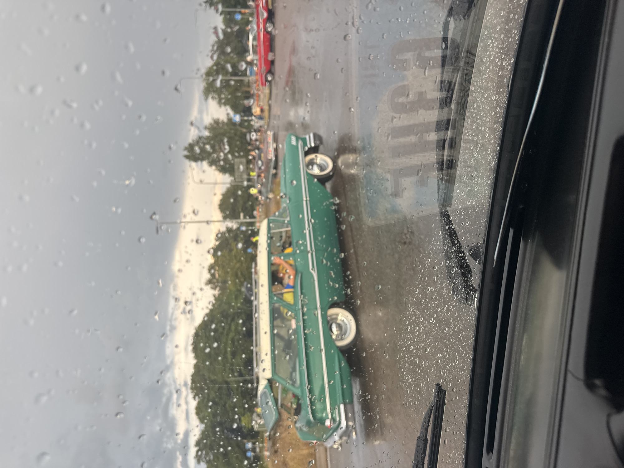 The image shows a rainy day scene, viewed from inside a car, likely in Falkenberg, Sweden. Water droplets are visible on the car's windshield, distorting the view of the outdoor environment. A vintage green station wagon with a white roof is the primary subject in the foreground, driving on a wet road. The car has whitewall tires and chrome accents. In the background, other classic cars, including a red one, are parked or driving, suggesting a car meet or event. Trees and streetlights are present, indicating an urban or suburban setting. The sky is overcast, consistent with rainy weather.