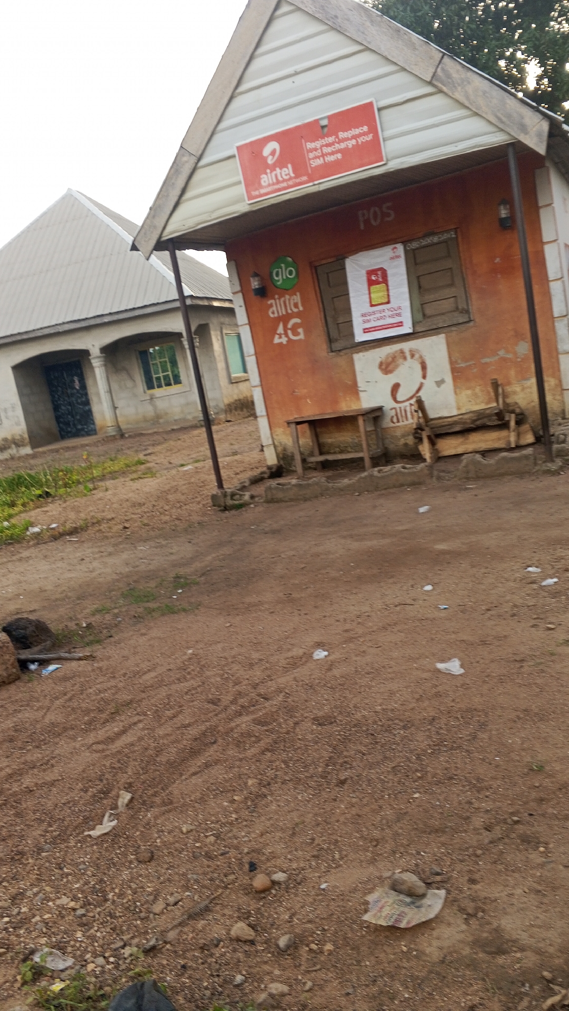 The image captures a small kiosk in Nyita, Nigeria, serving as a point of sale (POS) for telecommunication services. The kiosk is painted orange and white, with a corrugated metal roof. A red sign on the front advertises "Airtel" services, stating "Register, Replace and Recharge your SIM Here." Below this, another sign displays "glo" and "airtel 4G." A window on the front has a poster advertising "Register Your SIM Card Here." There are two wooden structures outside the kiosk, possibly for seating or display. The ground in front of the kiosk is dirt and gravel, with some scattered litter. In the background, a building with a light-colored roof and arched entrance is visible. The overall impression is of a rural or semi-urban commercial area.