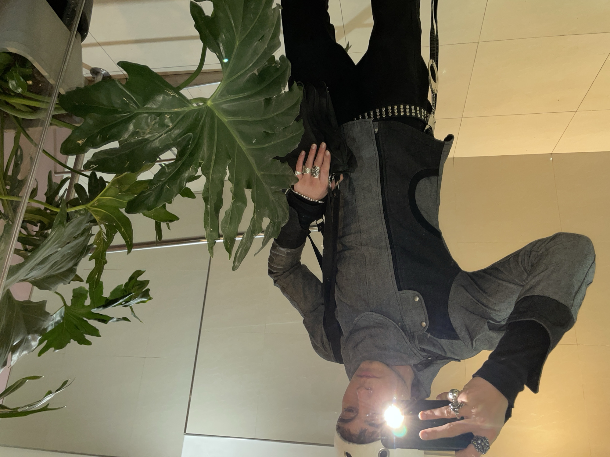 The image shows a reflection in what appears to be a mirror. A person is visible in the reflection, wearing dark clothing, a grey jacket, and a studded belt. They are holding a mobile phone and taking a photo. The reflection also captures indoor plants with large green leaves near the person. The background indicates a tiled wall and floor. The location is unknown, therefore I can't determine whether the picture was taken in Buenos Aires, Argentina.