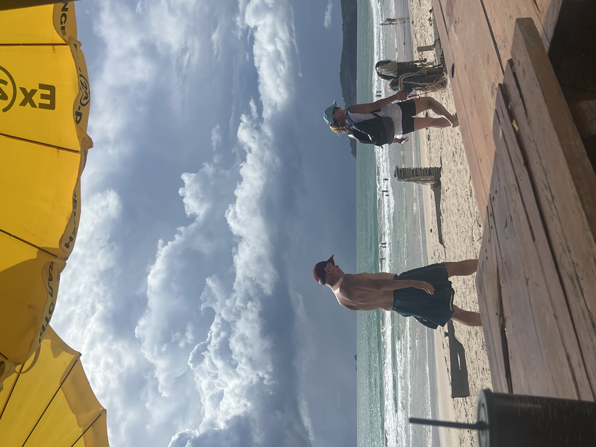 A medium-shot photograph, taken from under a partially visible yellow umbrella, shows a beach scene with a cloudy sky. In the foreground, a wooden picnic table is present. A black cup with a straw is on the left side of the table. Two people are walking on the sandy beach in front of the ocean.
On the left, a man wearing a red baseball cap and dark shorts walks towards the left, facing away from the camera. He is shirtless, revealing his back.
On the right, a woman wearing a light-colored top, dark shorts, and a light blue baseball cap walks towards the right, also facing away from the camera. She has a black backpack on.
The ocean has waves breaking on the shore, and a few other people are visible in the water and on the beach in the distance. Hills are visible on the horizon. The sky is filled with dramatic, dark clouds, suggesting an approaching storm. The location is identified as a beach in Choeng Thale, Thailand.