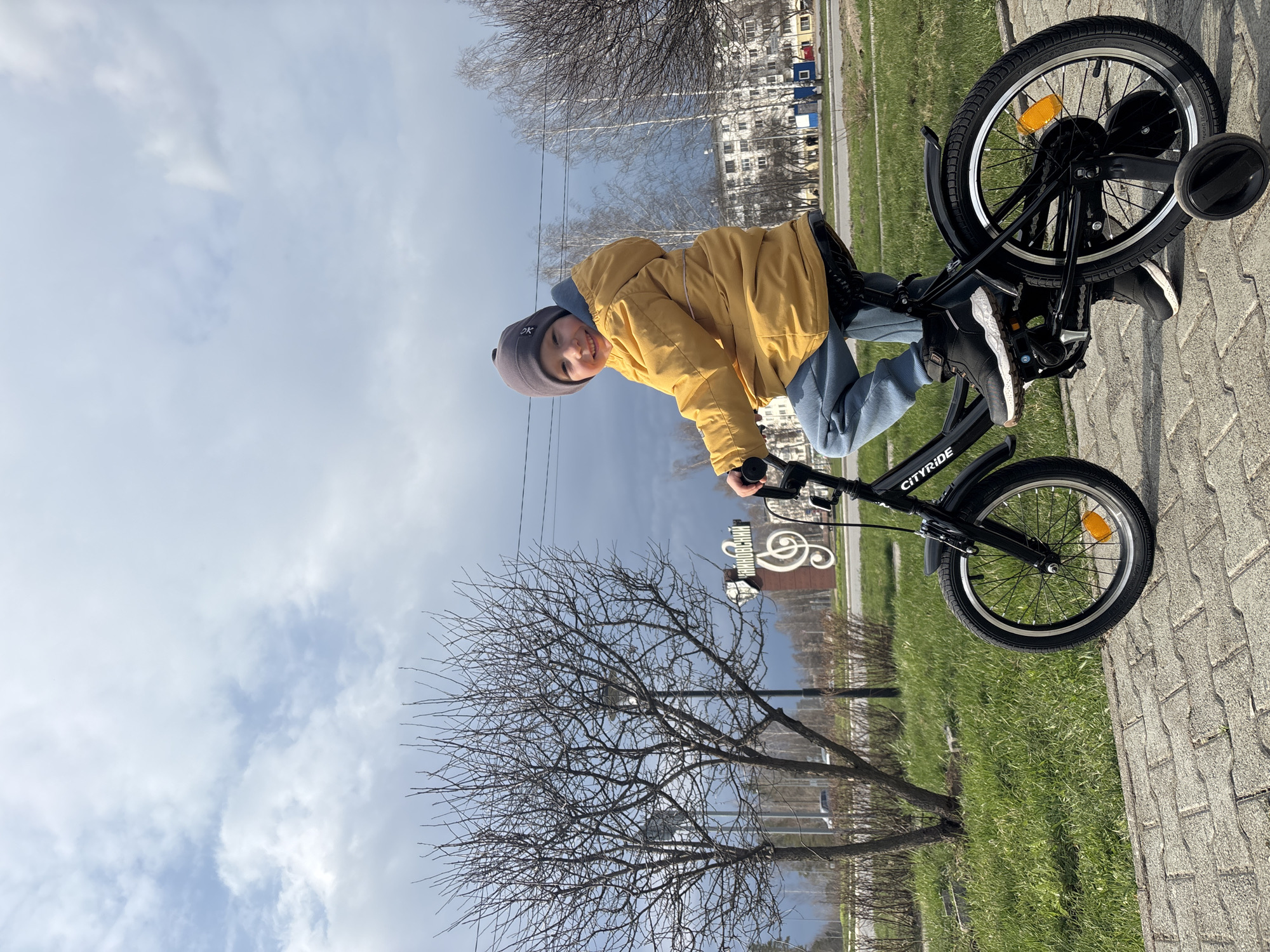 A young child, appearing happy and smiling, is seated on a black "CITYRIDE" bicycle equipped with training wheels. The child wears a yellow jacket, blue pants, and a grey beanie with the letters "OK" visible on it. Their feet are positioned on the bicycle's pedals, and hands are on the handlebars.
The setting is outdoors on a bright, partially cloudy day, likely during late winter or early spring in Chaikovsky, Russia. The ground beneath the child is a paved path made of grey interlocking pavers, with green grass lining its edge. Bare trees with intricate branches stand in the mid-ground and background, alongside distant residential buildings. A prominent landmark behind the child is a decorative sign or sculpture featuring a musical treble clef symbol and the Cyrillic text "ЧАЙКОВСКИЙ" (Chaikovsky), acknowledging the city's connection to the famous composer. The overall scene conveys a cheerful outdoor activity.