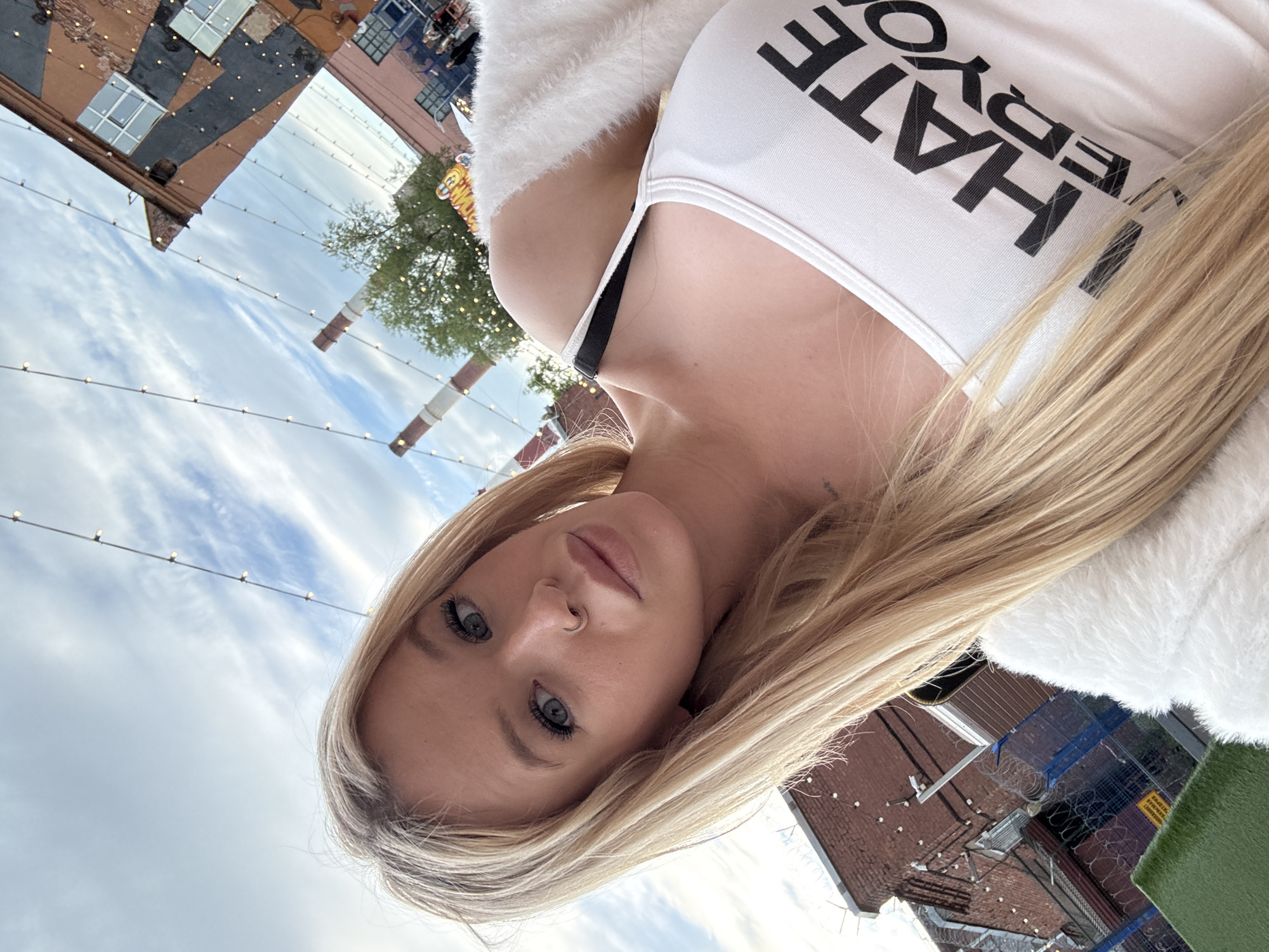 A young woman with blonde hair and blue eyes takes a selfie outdoors. She is wearing a white tank top with the words "HATE EVERYONE" printed on it in black, and a white fuzzy cardigan. The background shows an industrial area with brick buildings and smoking chimneys, characteristic of Saint Petersburg, Russia. String lights are visible across the top of the image. A chain-link fence and barbed wire are in the foreground, suggesting a possibly restricted or industrial zone. The sky is cloudy.
