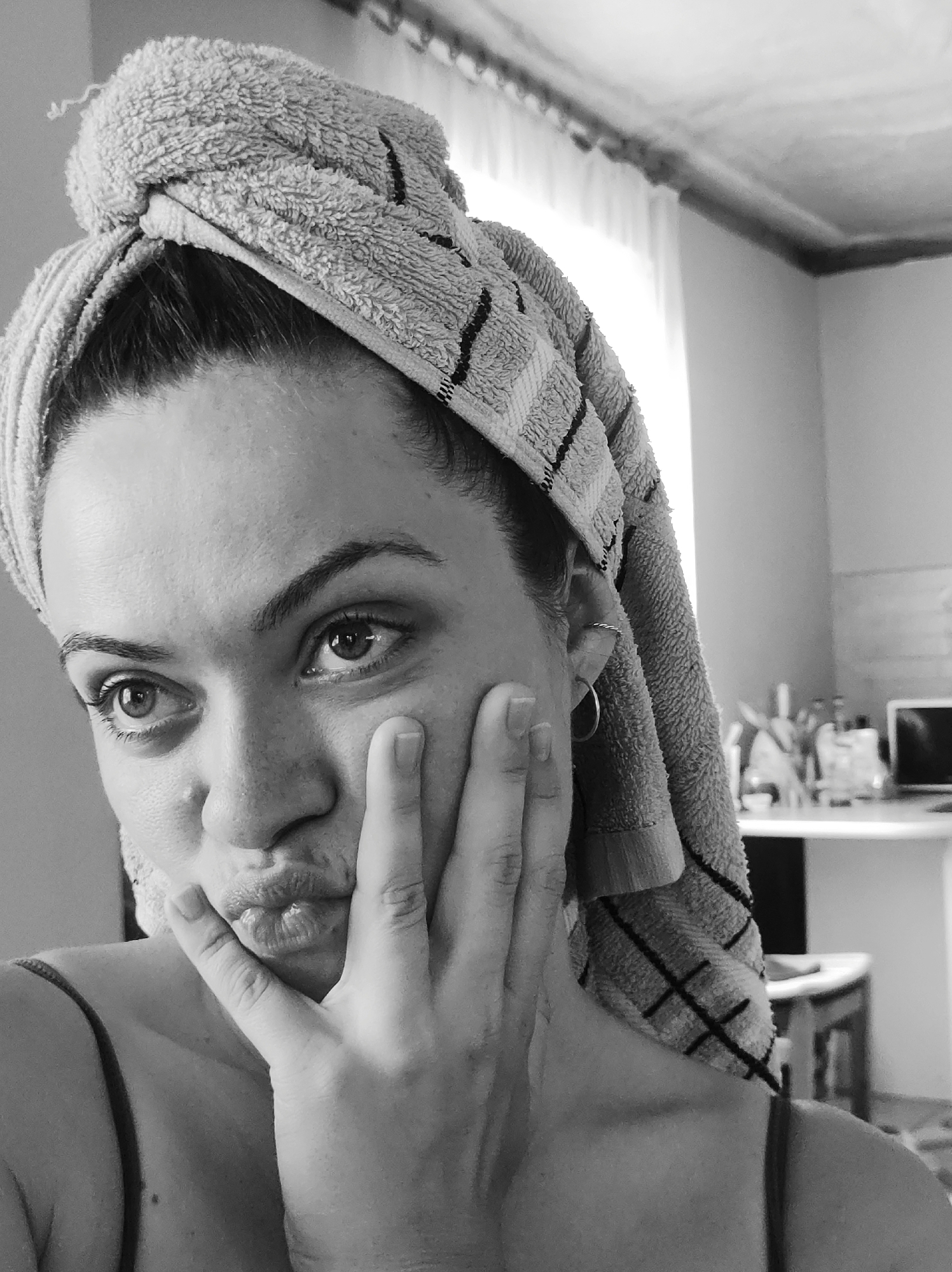 An indoor, black and white, eye-level photograph depicts a woman from the chest up, positioned slightly off-center to the left, within a room located in Unknown, Unknown. The woman, with dark hair, has her head wrapped in a light-colored, textured towel featuring dark striped patterns. A loose thread protrudes from the top of the towel. She is looking towards the left side of the frame with a direct gaze. Her lips are pursed, forming a "kiss" or "duck" face expression, and her right hand is placed against her left cheek, with fingers slightly splayed. She wears a simple top with a thin strap visible on her right shoulder. Two small earrings are present in her visible left ear: a circular hoop in the lobe and a twisted ring in the helix. The background is out of focus, revealing a bright light source, likely a window, to the upper right. Above the window, a ceiling with decorative molding is visible. Further to the right, a blurred table or counter holds several indistinct objects, including what appears to be a small screen or monitor.