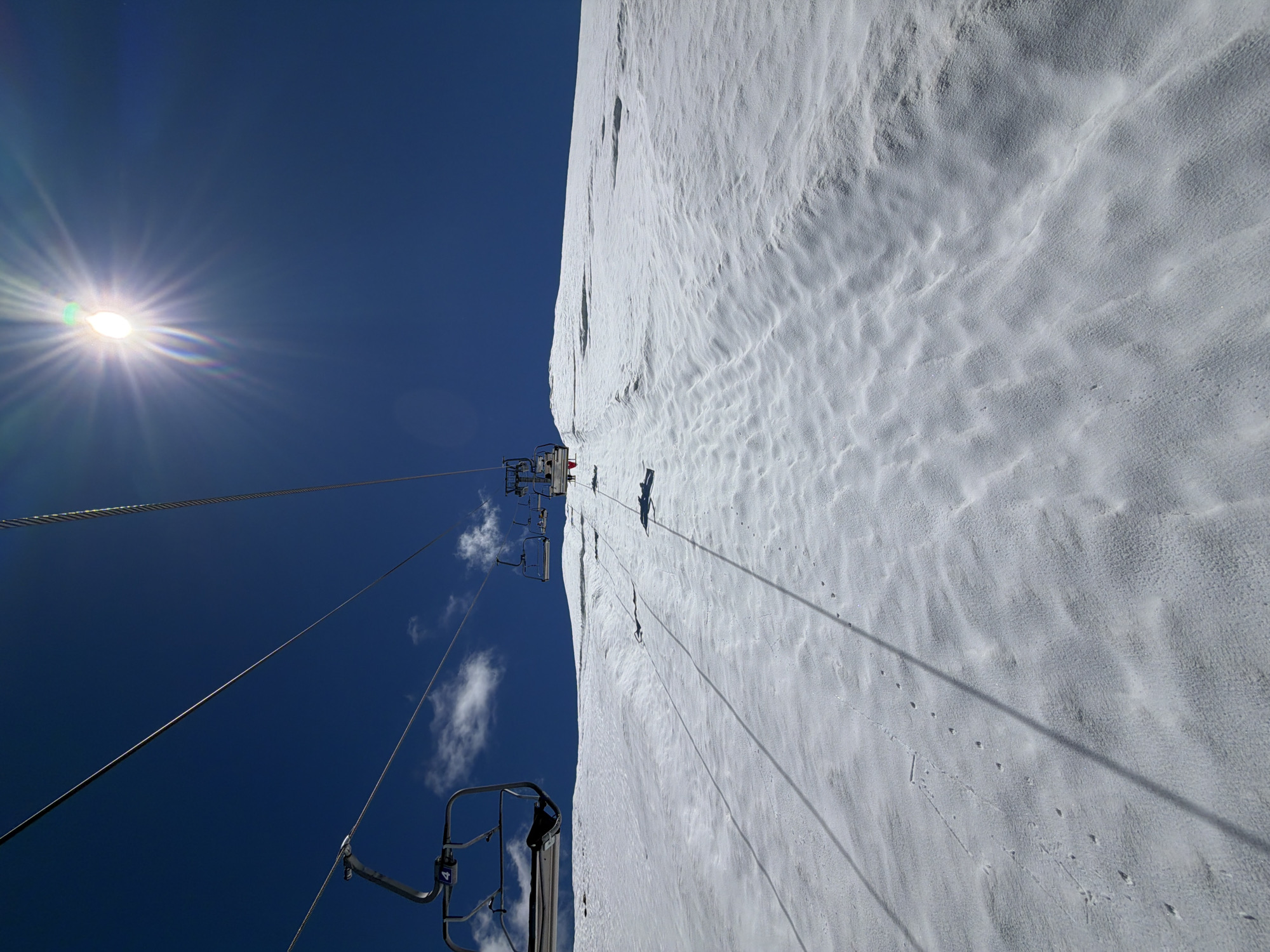 The image shows a ski lift with multiple chairs ascending a snow-covered slope under a bright blue sky with the sun prominently featured. The snow surface has wind-blown textures and appears to be a ski slope. Several chairlifts are visible, moving upwards. The scene is outdoors, likely at a ski resort in the mountains. The location is identified as Verkhne-Teberdinskoye Rural Settlement, Russia, which is known for its mountainous terrain and ski resorts. There are no people visible in the chairs or on the slope. The sun is high in the sky, casting shadows and creating lens flare.