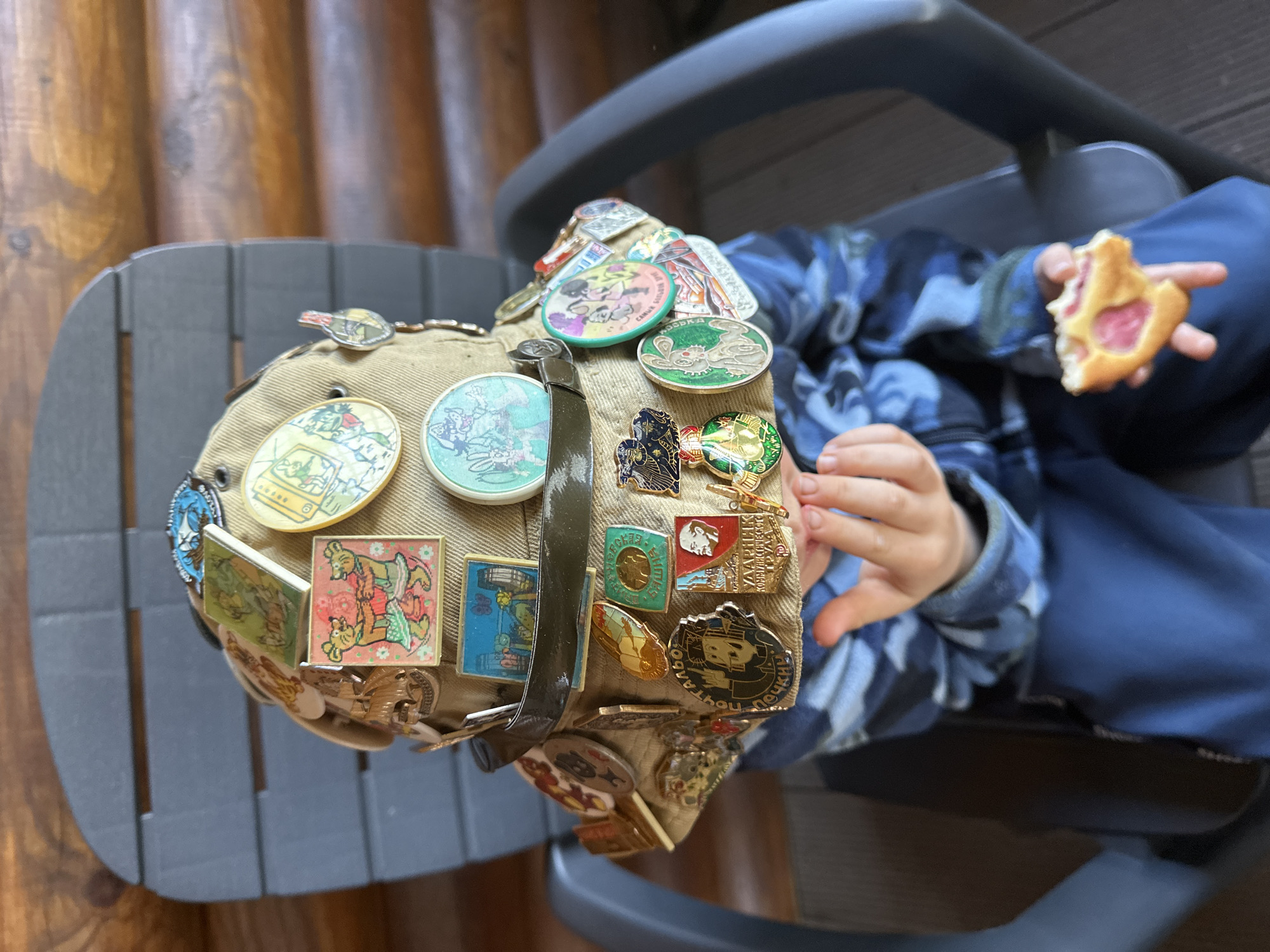 A child wearing a blue camouflage-patterned shirt sits in a grey plastic chair, holding a pastry. The child is positioned indoors near a wall made of brown wooden logs. The focus is a beige cap densely covered in pins and badges. The pins appear to be vintage or collectible. Although some pins have Russian writing on them and I believe this picture was taken in Russia, there is no explicit evidence of the city of Nizhny Novgorod in this picture.