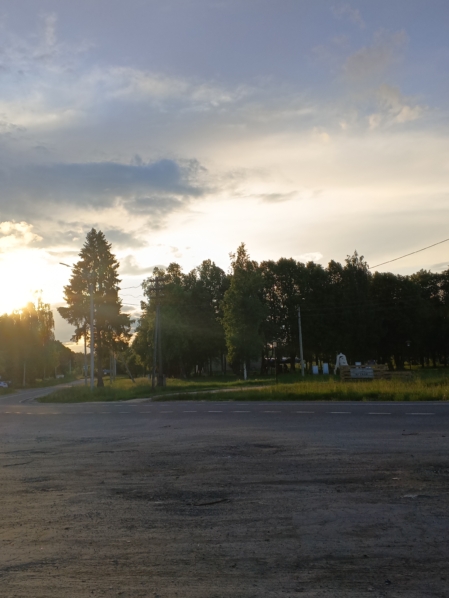 This image depicts a rural or semi-rural landscape scene in Красный Маяк, Russia, captured during the late afternoon or sunset. The sun, positioned low on the left, creates a strong golden light and a significant lens flare, illuminating the clouds and the distant horizon. The sky features a mix of soft blue and scattered clouds, transitioning to a bright, hazy white and golden hue where the sun is brightest.
In the foreground, a paved road with dashed white lane markings stretches across the frame, leading towards a gentle curve on the left. The road surface appears dark and somewhat textured. To the right of the road, a strip of green grass precedes a dense treeline, which forms the mid-ground and background. This treeline consists of various trees, including a prominent tall coniferous tree (likely a spruce) towards the left, and numerous deciduous trees. Utility poles with power lines are visible along the edge of the treeline, adding an element of human infrastructure to the natural setting.
Further to the right, amidst the grass and in front of the trees, a small, light-colored structure, possibly a temporary tent or stall, is visible. It rests upon a base of wooden pallets. On one of these pallets, the Cyrillic word "ДРУЖБА" is clearly discernible, which translates to "Friendship" in English. There are no people visible, and no specific activity is taking place, contributing to a serene and tranquil atmosphere under the golden hour light.