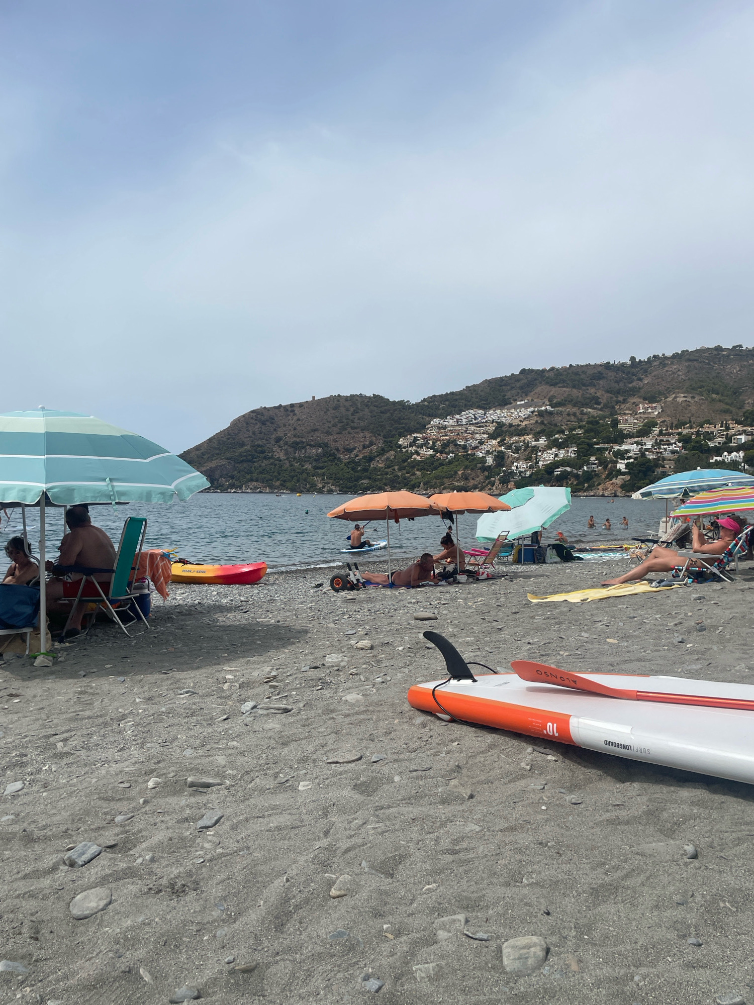 The image depicts a sun-drenched beach scene in La Herradura, Spain. The foreground is dominated by a white and orange paddleboard with a black fin and paddle resting on the grey, pebble-strewn sand. In the mid-ground, several beach umbrellas, primarily in blue and orange, provide shade for sunbathers. People are seen relaxing on chairs, lying on towels, and swimming in the calm, blue sea. A brightly colored kayak rests on the shore. The background reveals a steep, verdant hillside dotted with white buildings, characteristic of the coastal towns in the region, and the clear sky above. The overall atmosphere is one of leisure and relaxation on a warm day.