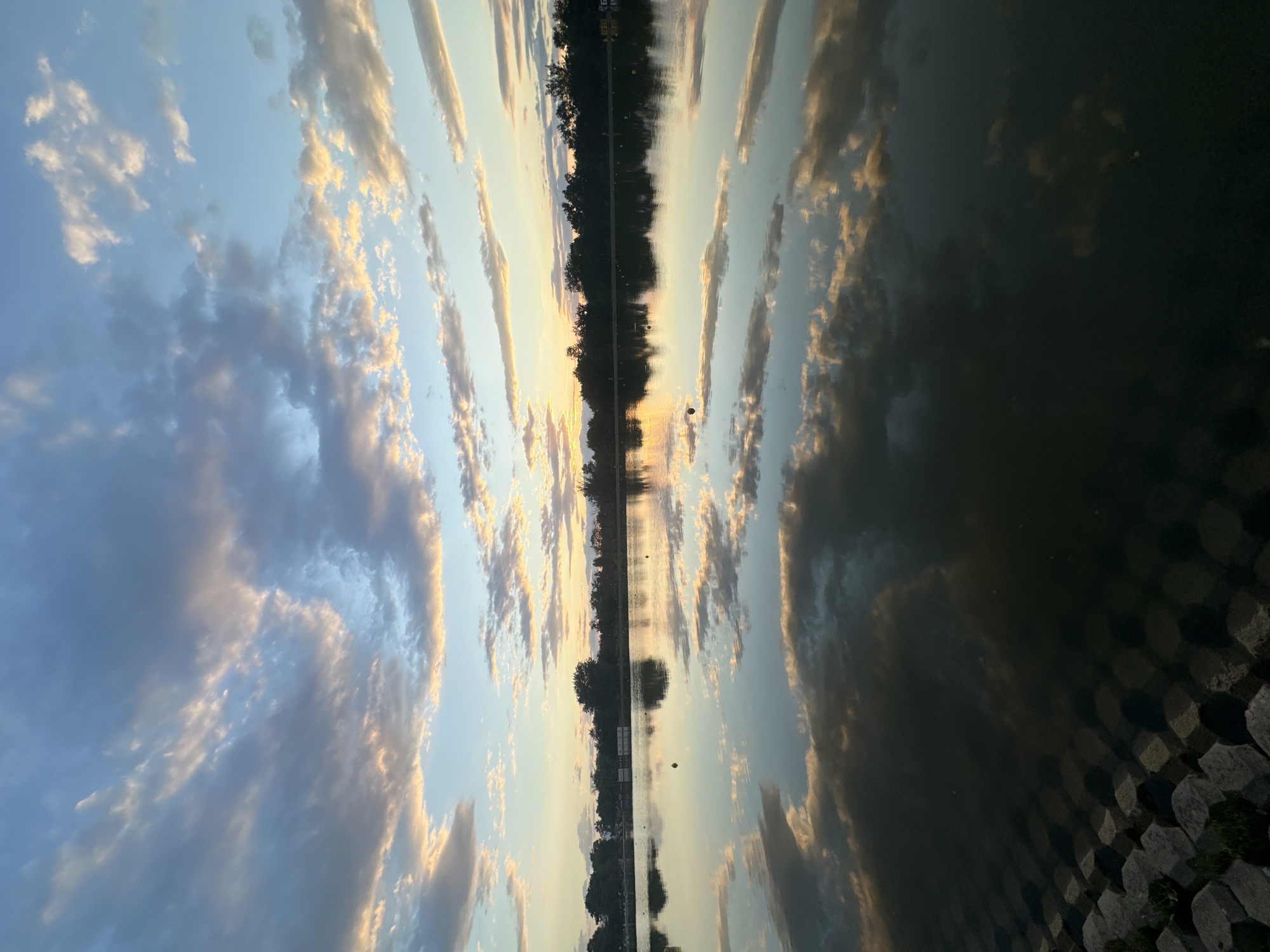 This is a vertical, eye-level, medium shot of a lake reflecting the sky and trees at sunset. The sky is filled with scattered clouds, tinged with orange and pink from the setting sun. The water is calm, creating a mirror image of the sky and the tree line on the far bank. The foreground shows a textured stone embankment leading into the water. The scene appears to be in a park or recreational area.