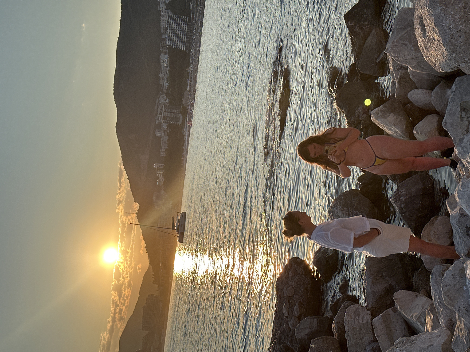 At sunset in Rafailovići, Montenegro, two women stand on a rocky shoreline. The woman on the left wears a white shirt and light-colored shorts, while the woman on the right is in a yellow bikini. The sun, partially obscured by clouds, casts a golden glow on the water, reflecting on the surface and illuminating a sailboat in the distance. Buildings and hills line the coast behind the water, suggesting a populated area. The overall scene captures a serene moment at dusk by the Adriatic Sea.