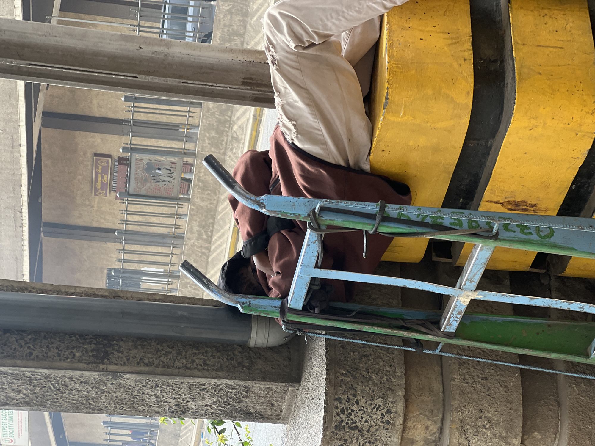 A street scene, taken in Nairobi, Kenya, shows a person working on an elevated scaffold above a building. The person wears light-colored pants with distressed hems, a brown jacket, and black gloves. The scaffold is painted in shades of blue and green. Below the scaffold is a concrete structure with a pipe running along its surface. In the background, there is a building with metal bars on the windows, as well as a sign with text. The environment appears to be urban and potentially congested.