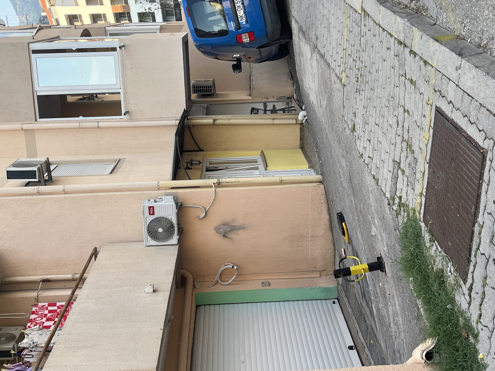 The image shows a narrow alleyway between buildings with a paved ground. On the left side, there is a multi-story building with beige and peach-colored walls. Windows and air conditioning units are visible on the building. One window, with a white frame, is open, revealing the interior. Below the open window, there is a smaller open grate. Further down, a white rolling door is partially visible. On the right side of the alley, there is a gray paved surface with some grass growing through the cracks. A metal grate is set into the ground. In the background, a blue car with a license plate that reads "NK DK51" is parked. Yellow and black traffic control devices are present on the paved surface. The scene appears to be an urban environment. The lighting suggests it is daytime, possibly late afternoon or early evening, with soft shadows. There are no people visible in the image. The overall impression is of a utilitarian urban space, possibly a service alley or a residential courtyard.