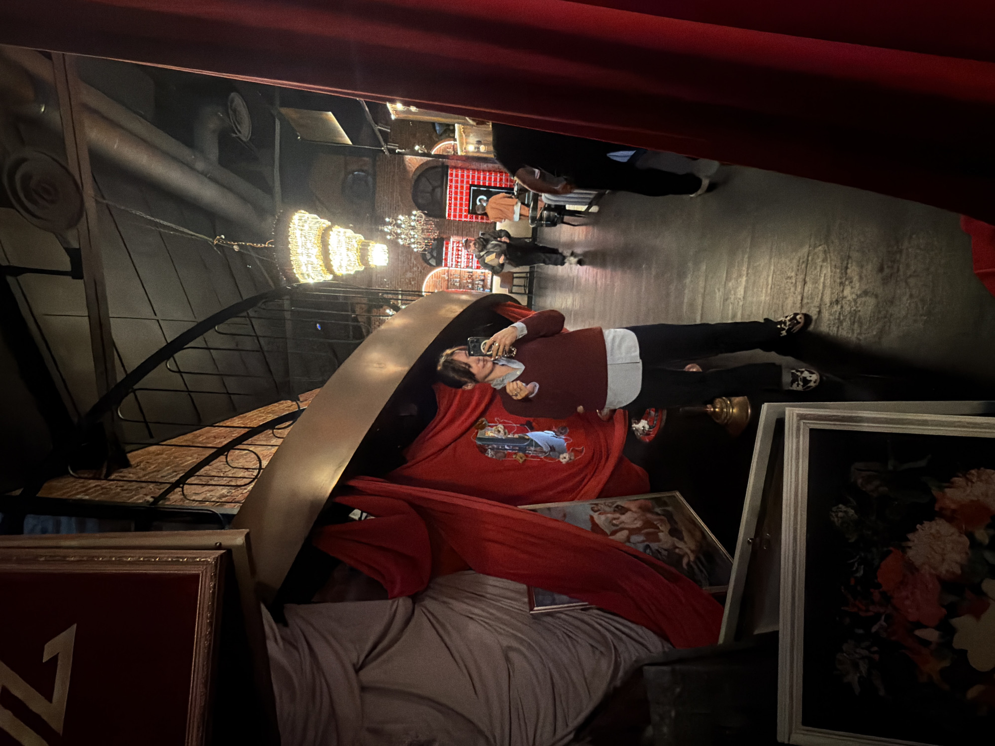 The scene is an indoor space, likely a restaurant or bar, in Moscow, Russia. The perspective is slightly skewed, looking across the space. Several people are present, including an individual in the foreground holding a phone, possibly taking a photo. The decor is ornate with chandeliers hanging from the ceiling. There are architectural details such as exposed ventilation and brickwork visible. Various objects are scattered around including picture frames, draped fabric and furniture, potentially related to a theater stage or a decorated display. The overall impression is of a theatrical or stylized setting.