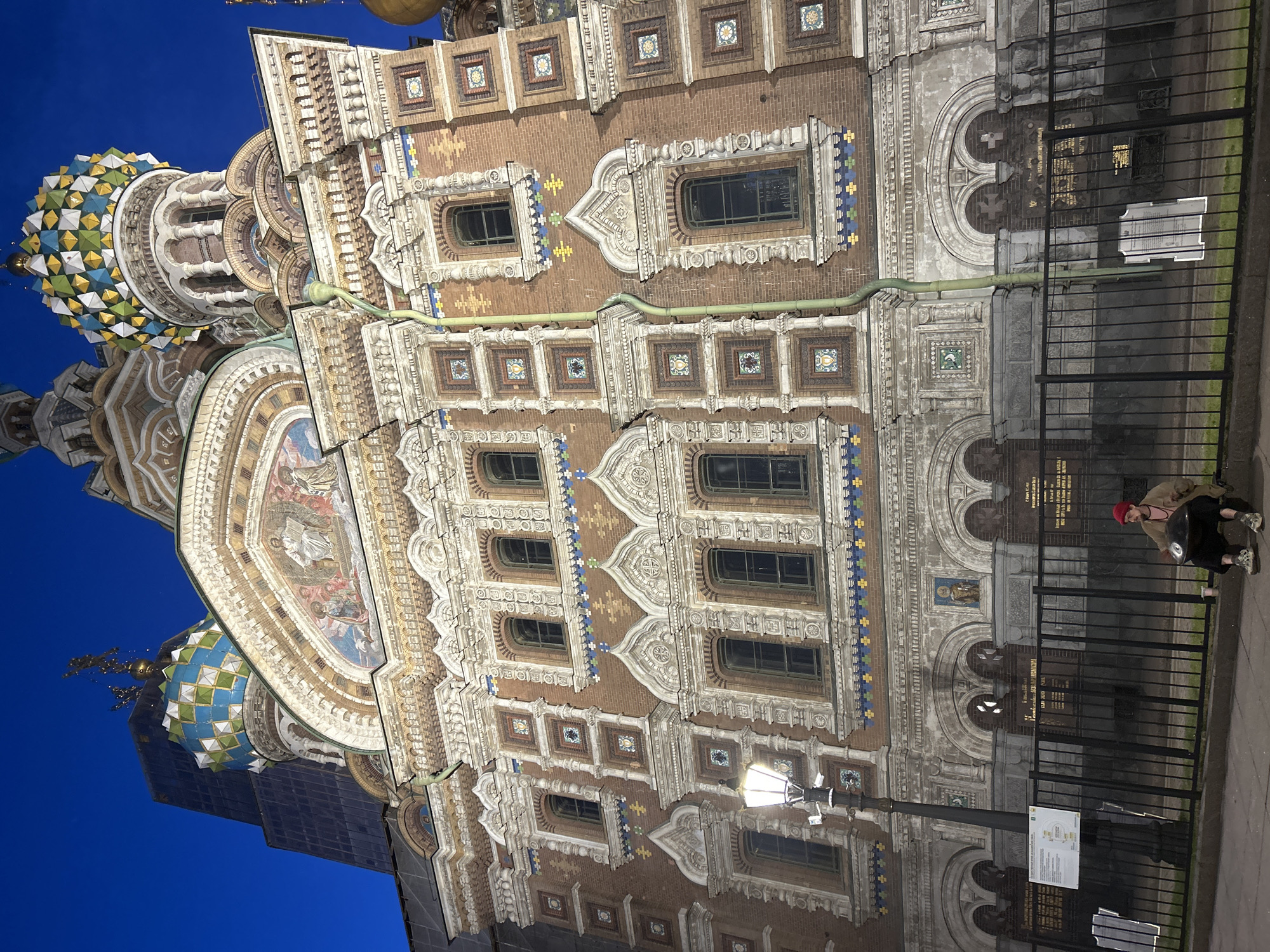 The image shows the Church of the Savior on Spilled Blood in St. Petersburg, Russia at dusk. The ornate brick building features colorful onion domes and intricate mosaics. In the foreground, a person sits on the sidewalk behind a black metal fence, playing a handpan. The person is wearing a red hat, a light-colored jacket, and dark pants. A street lamp stands to the left of the frame. The sky is a deep blue, indicating the time is after sunset.