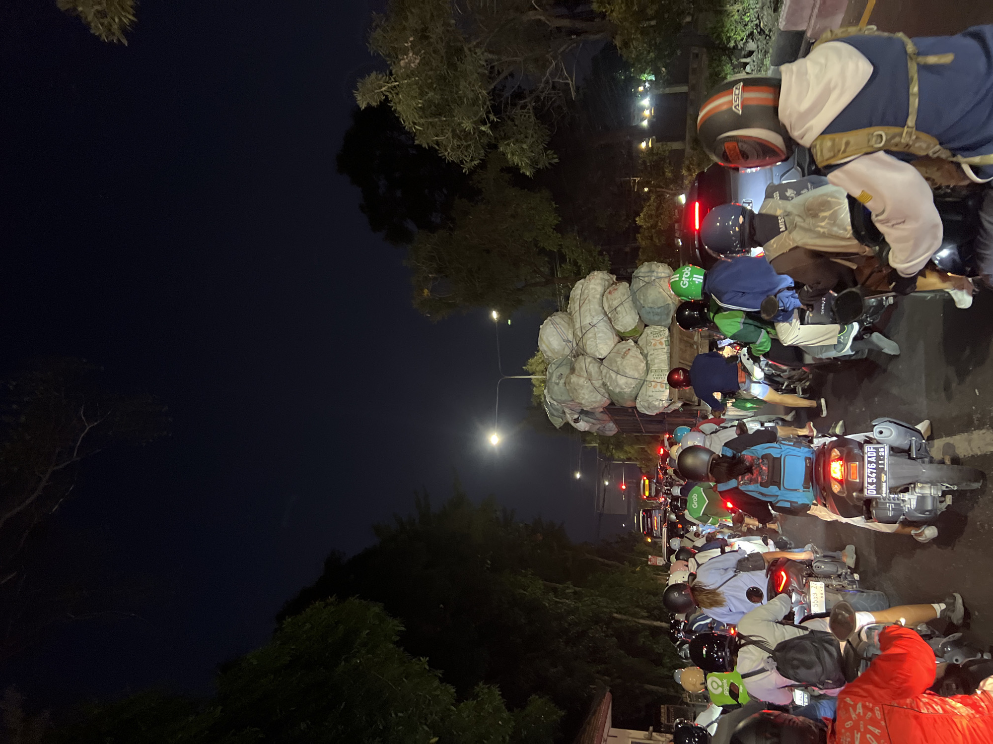 The image captures a nighttime traffic jam on a street in Denpasar, Indonesia, likely in an urban or suburban area based on the density of vehicles and surrounding trees. The scene is dominated by a long line of motorcycles, with some cars and a truck visible further down the road. Many of the motorcyclists are wearing helmets and some are adorned with green and blue jackets, suggesting they might be ride-sharing service drivers, possibly from "Grab," a prominent platform in Southeast Asia. The truck in the foreground is heavily laden with large, white, cylindrical bales, possibly agricultural products or waste, secured with netting. The street is illuminated by streetlights and the headlights and taillights of the vehicles, creating a somewhat dim and atmospheric setting. The presence of lush trees lining the street indicates a tropical environment, consistent with Denpasar. The overall impression is one of slow-moving, congested traffic in the evening.