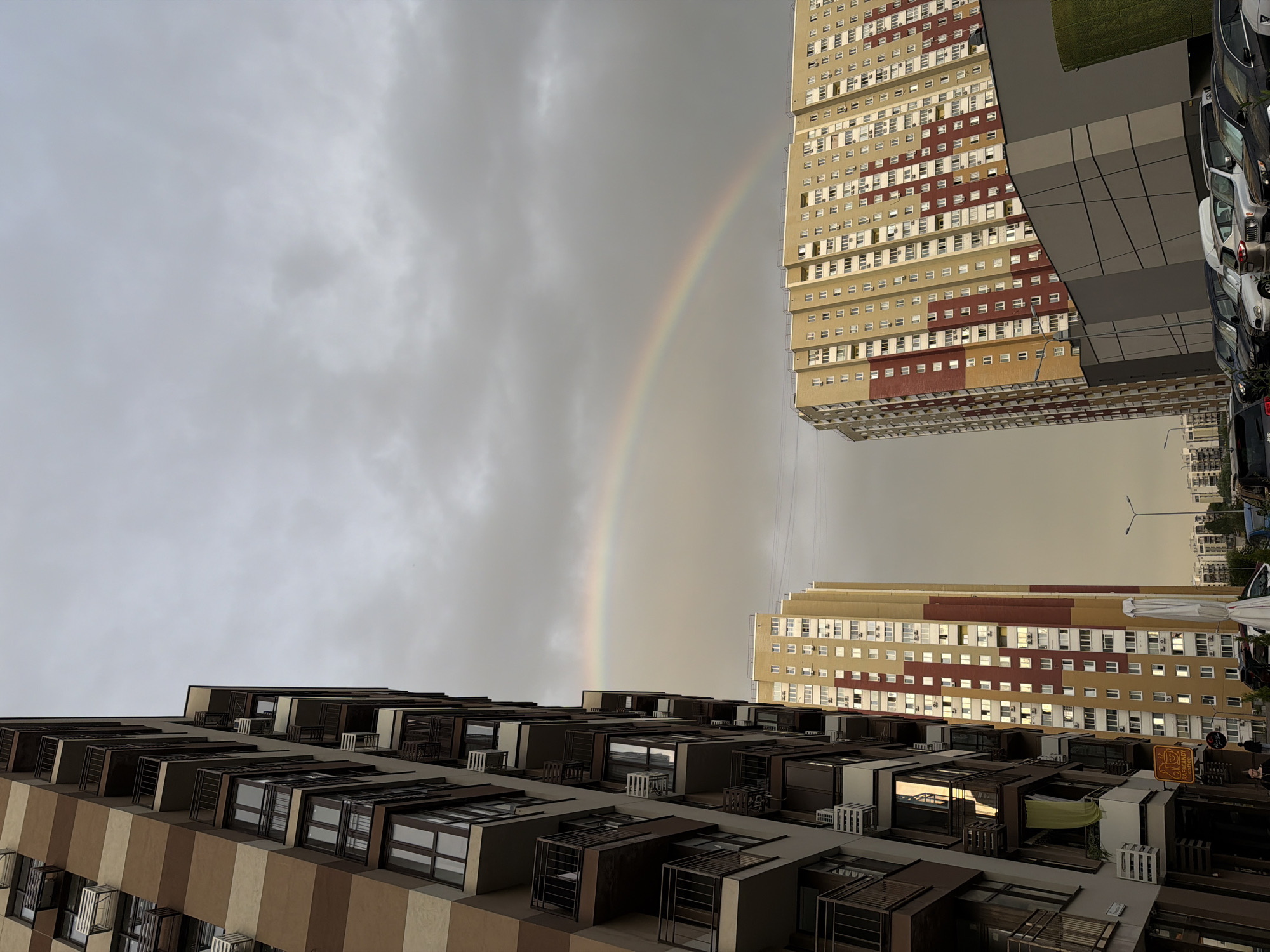 The image shows a view of apartment buildings with a rainbow in the sky, taken in Kyiv, Ukraine. On the left, a tall apartment building with a striped facade of brown and beige colors occupies the foreground. The building features multiple balconies with dark railings and numerous windows. To the right, two other apartment buildings are visible. The taller one in the middle has a yellow and red facade with many windows. In front of it, a modern building with gray and metallic paneling is visible. The sky is overcast with clouds, and a faint rainbow arcs across the scene. Several cars are parked in the foreground, and a lamppost stands near the right edge of the frame. The overall impression is a somewhat subdued urban landscape under a dramatic sky.