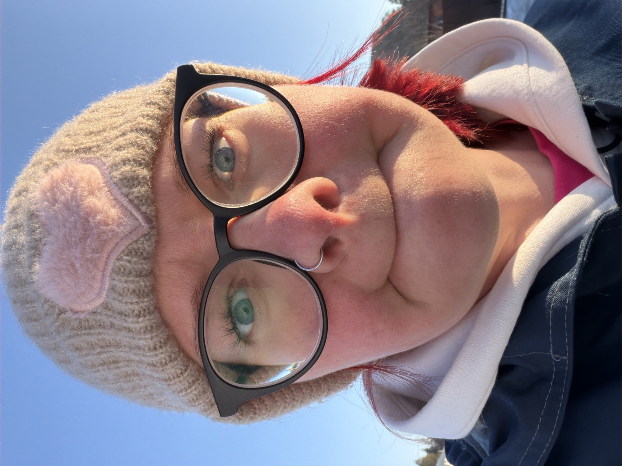 This image is a close-up, front-facing portrait of an individual located outdoors in Chaikovsky, Russia. The subject, likely female, has fair skin with a flushed appearance on the cheeks and nose. They wear a light beige knitted beanie featuring a pink, textured heart-shaped appliqué on the left front. Black-rimmed eyeglasses are worn, framing light-colored eyes, with the left eye appearing green and the right eye appearing blue. A silver hoop nose ring is present in the left nostril. Sections of red-dyed hair are visible around the ears and neck. The individual's lips are pressed together, creating a puckered expression. Attire includes a white hooded garment layered beneath a navy blue outer jacket, with a pink undergarment visible at the neckline. The scene is illuminated by direct sunlight from the right, casting shadows on the left side of the subject's nose and beneath the lower lip, indicating a low sun angle. The background consists of a clear blue sky, with indistinct dark vegetation and a faint building structure visible towards the lower right, suggesting an outdoor, potentially cold environment.