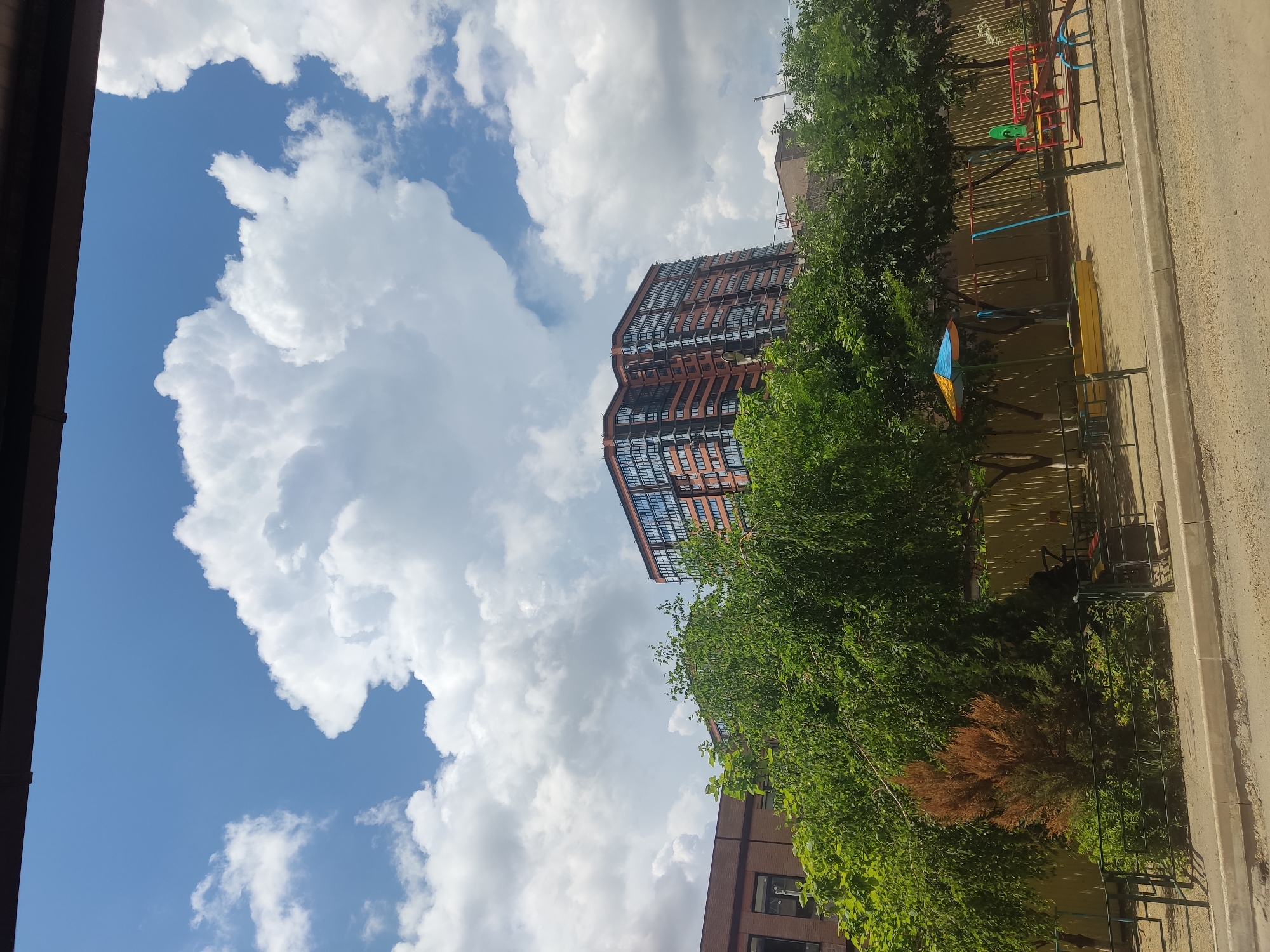 The image shows a cityscape in Krasnodar, Russia, featuring a modern, multi-story building with a distinct architectural design, surrounded by lush green trees and foliage. To the right of the building is a playground with colorful play structures. The sky is partially covered with fluffy white clouds, casting shadows across the scene. The photograph appears to have been taken from a low angle, looking upwards towards the building and sky. The composition suggests a perspective from street level or a nearby building.