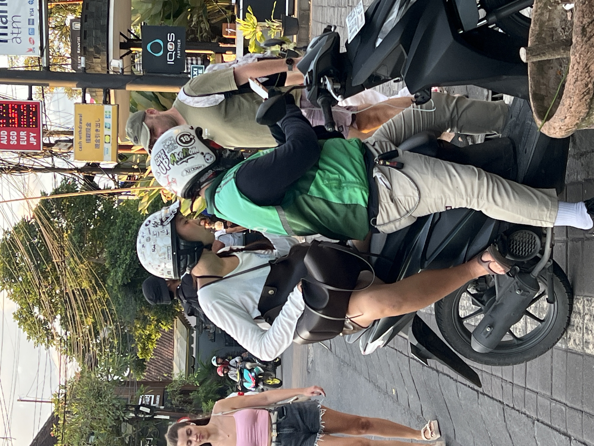 Here is a description of the image:
The image shows a street scene in Ubud District, Indonesia. Three people are on motorbikes, which appear to be scooters. The drivers of two scooters are wearing helmets. One of the drivers wears a green vest. Another driver is wearing a light green shirt, tan pants, and a hat. A woman stands on the sidewalk; she is wearing a pink top and denim shorts. Overhead signs provide information on ATMs and currency exchange rates. The OCR detected signs indicate ATMs are available and provide currency exchange rates. Additionally, a sign for IQOS is visible.