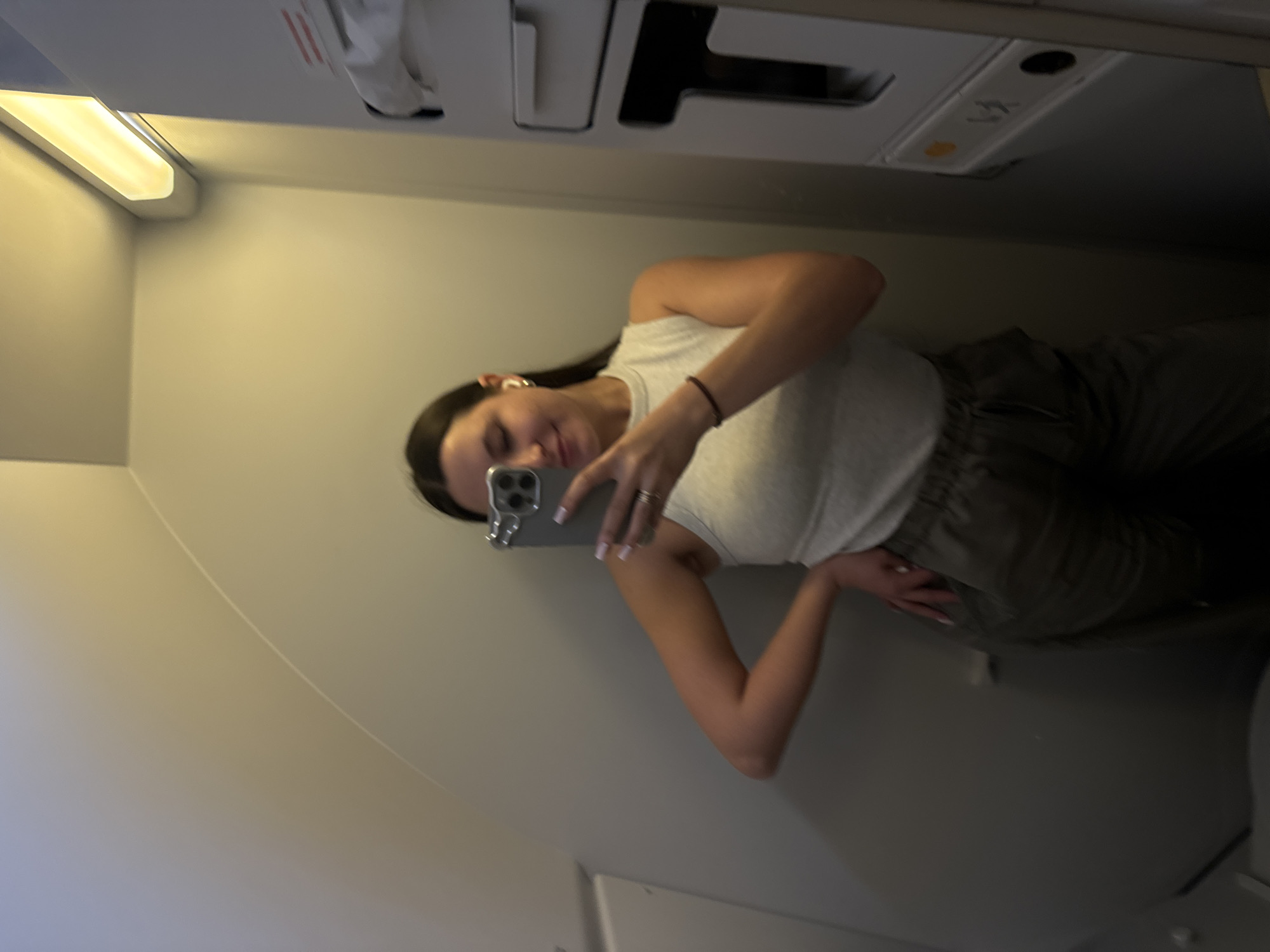 A woman is taking a selfie in what appears to be the lavatory of an airplane. She is wearing a light-colored tank top and dark pants. The airplane lavatory has standard features such as an overhead light, air vents, and a compartment with a disposal bin. The location is not Moscow, Russia.