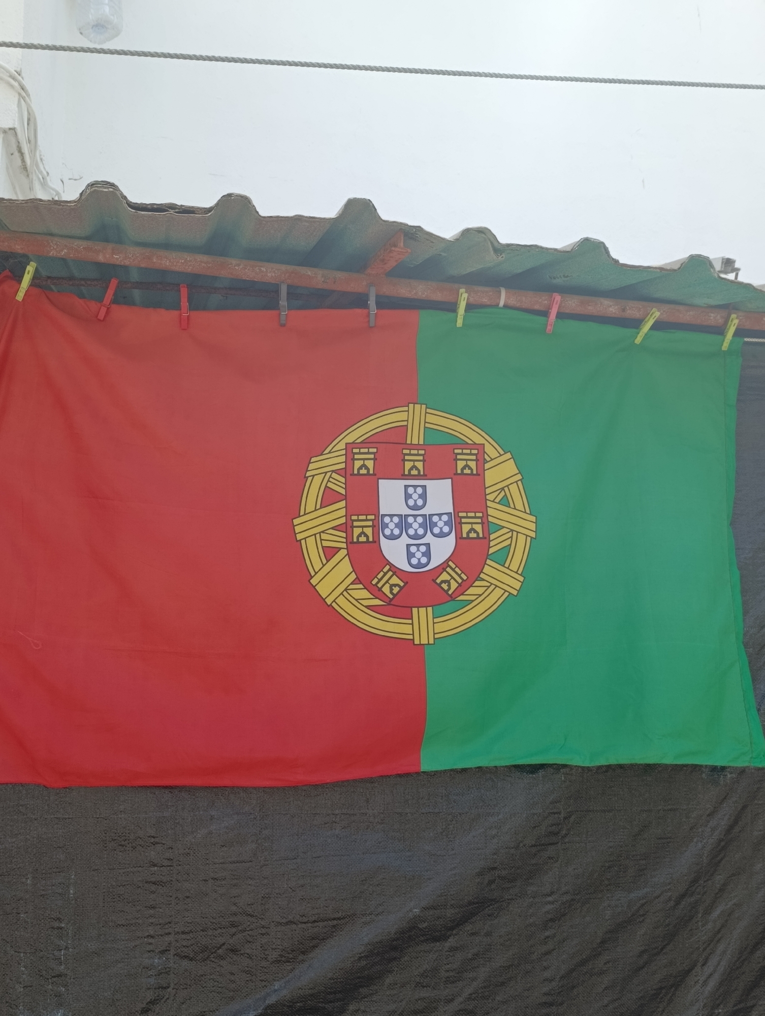 The image depicts the flag of Portugal hanging outdoors. The flag is affixed to a rusty metal pipe with several colored clothespins. The flag is divided vertically into red and green sections, with the national coat of arms centered on the dividing line. The coat of arms features a shield with five smaller shields and a castle emblem. Behind the flag, a corrugated roof structure is visible, supported by a reddish-brown metal beam. A taut rope runs horizontally across the top of the frame, and a partially visible plastic bottle is present in the upper left corner. The bottom of the image is covered by a black tarp-like material. The scene appears to be in an outdoor setting, likely a residential area, given the presence of laundry line and common household items. The location of the city of Setúbal, Portugal is not discernable from the visual elements present in the image.