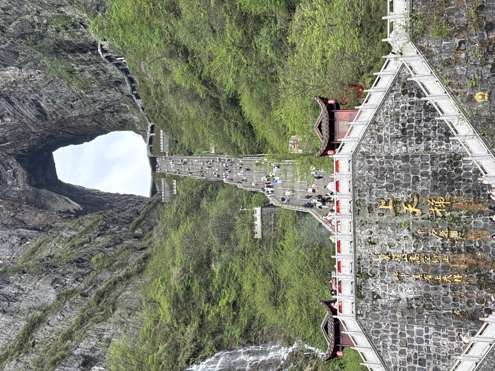 This image depicts Tianmen Cave, also known as Heaven's Gate, in the city of Zhangjiajie, China. The cave is a large natural archway through a mountain. A long staircase with many people ascending it leads from the foreground toward the cave opening in the background. Lush green vegetation covers the surrounding cliffs. A stone structure with traditional Chinese architectural elements and inscriptions stands near the base of the staircase. There are waterfalls on the sides of the path. The sky is visible through the cave opening.