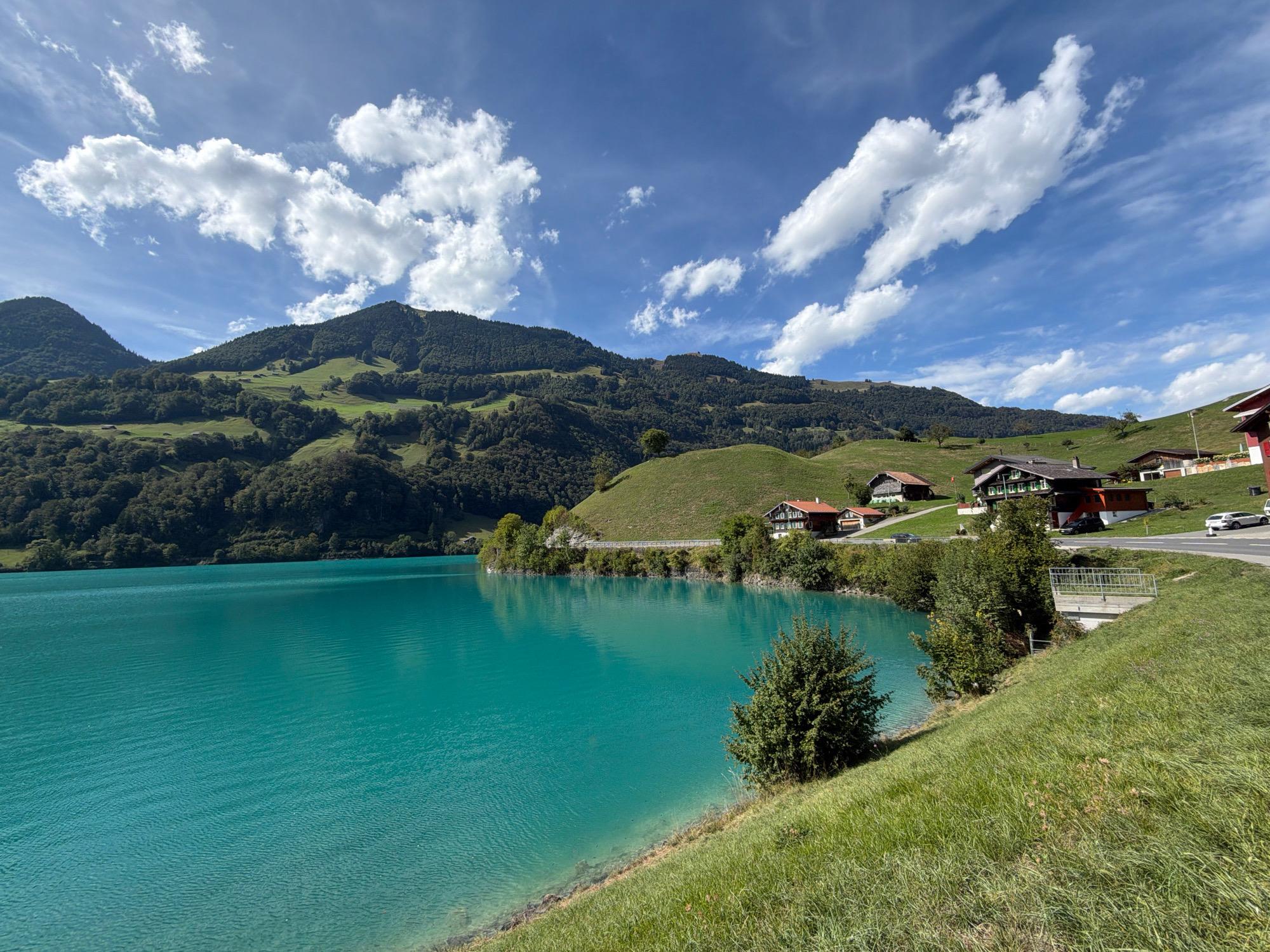 The image shows a scenic view of a turquoise lake bordered by lush green hills and mountains under a bright blue sky with scattered white clouds. Several traditional Swiss-style houses with red roofs are nestled on the slopes overlooking the lake. A paved road winds along the shoreline, with a few cars visible. The foreground features a grassy slope leading down to the clear water. The environment appears serene and rural, likely in a mountainous region known for its natural beauty.