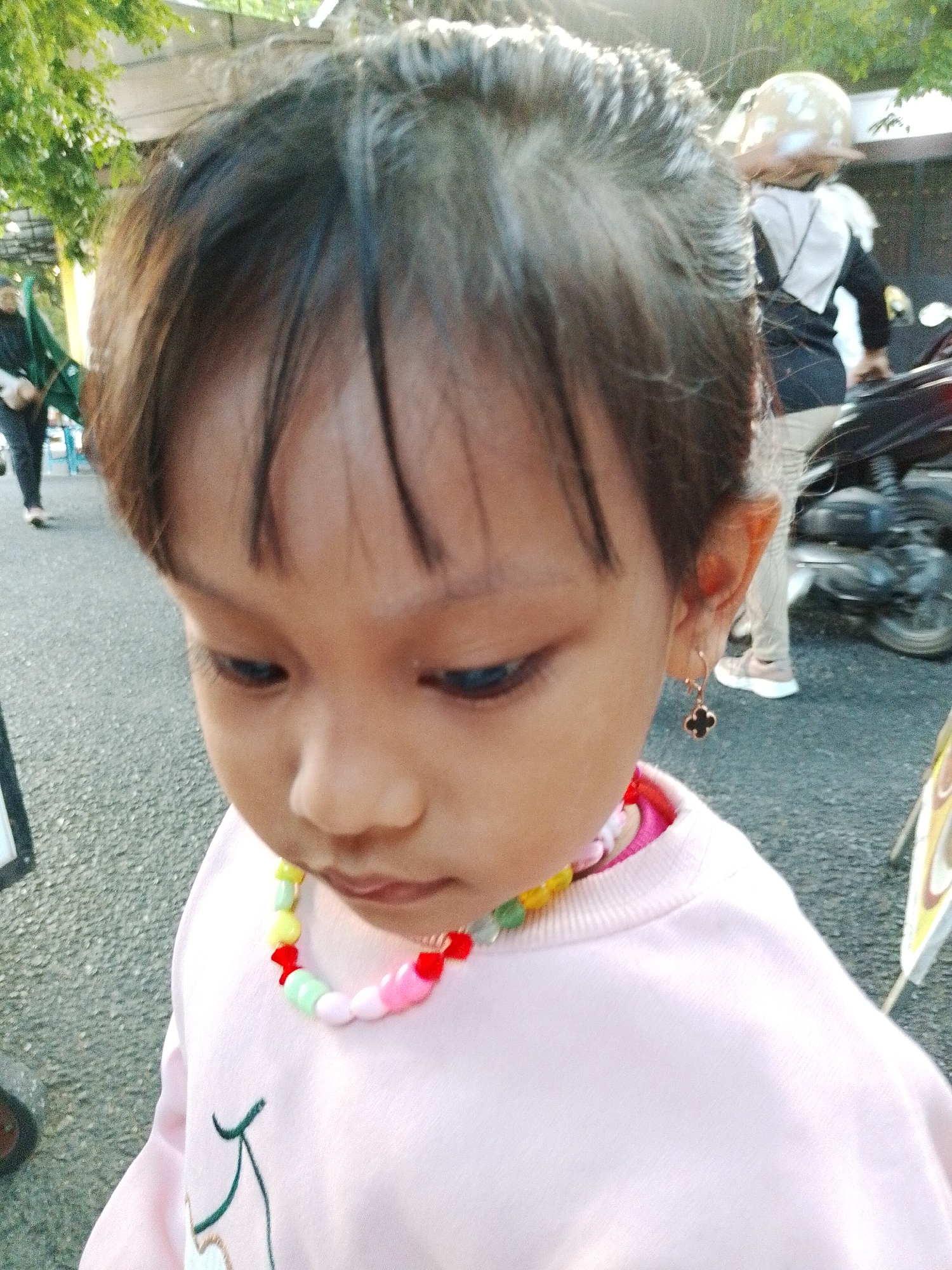 The image captures a close-up of a young girl wearing a pink sweatshirt and a colorful beaded necklace. She has dark hair and is looking downwards. On her right ear, she wears a clover-shaped earring. In the background, there are hints of an outdoor setting, possibly in Kecamatan Pati, Indonesia, with a motorcycle and other people visible, suggesting a public space like a street or market. The lighting indicates it is daytime.
