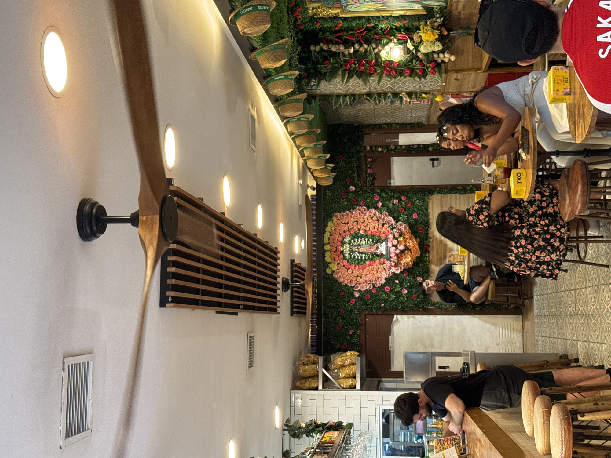 The image shows the interior of a restaurant in Valencia, Spain. A large wooden ceiling fan with black blades is prominently featured, along with recessed ceiling lights and a ventilation grate. The walls are decorated with artificial greenery, floral arrangements, and straw hats.
In the foreground, a man with dark, curly hair is standing at a wooden bar, leaning on it and looking down. He is wearing a black t-shirt and shorts. Several round, cushioned stools are arranged at the bar.
Further inside the restaurant, several patrons are seated at tables. A man with dark hair is sitting at a small table, looking at his phone. A woman with long, dark hair and a floral dress is seated at another table, facing away from the camera. Two women are seated at a table in the middle ground; one with long, dark curly hair is holding a piece of food to her mouth, and the other with blonde hair is looking at her. Several small yellow boxes with text are on the tables.
In the background, bags of snacks are stacked on shelves, and a decorative floral wreath with an image of the Virgin Mary is displayed on a green wall. The floor is tiled with a geometric pattern. A large, white refrigerator or cooler is visible behind the bar. A sign with the text "TKO" is on one of the tables. Another sign, partially visible on the right, has red lettering.