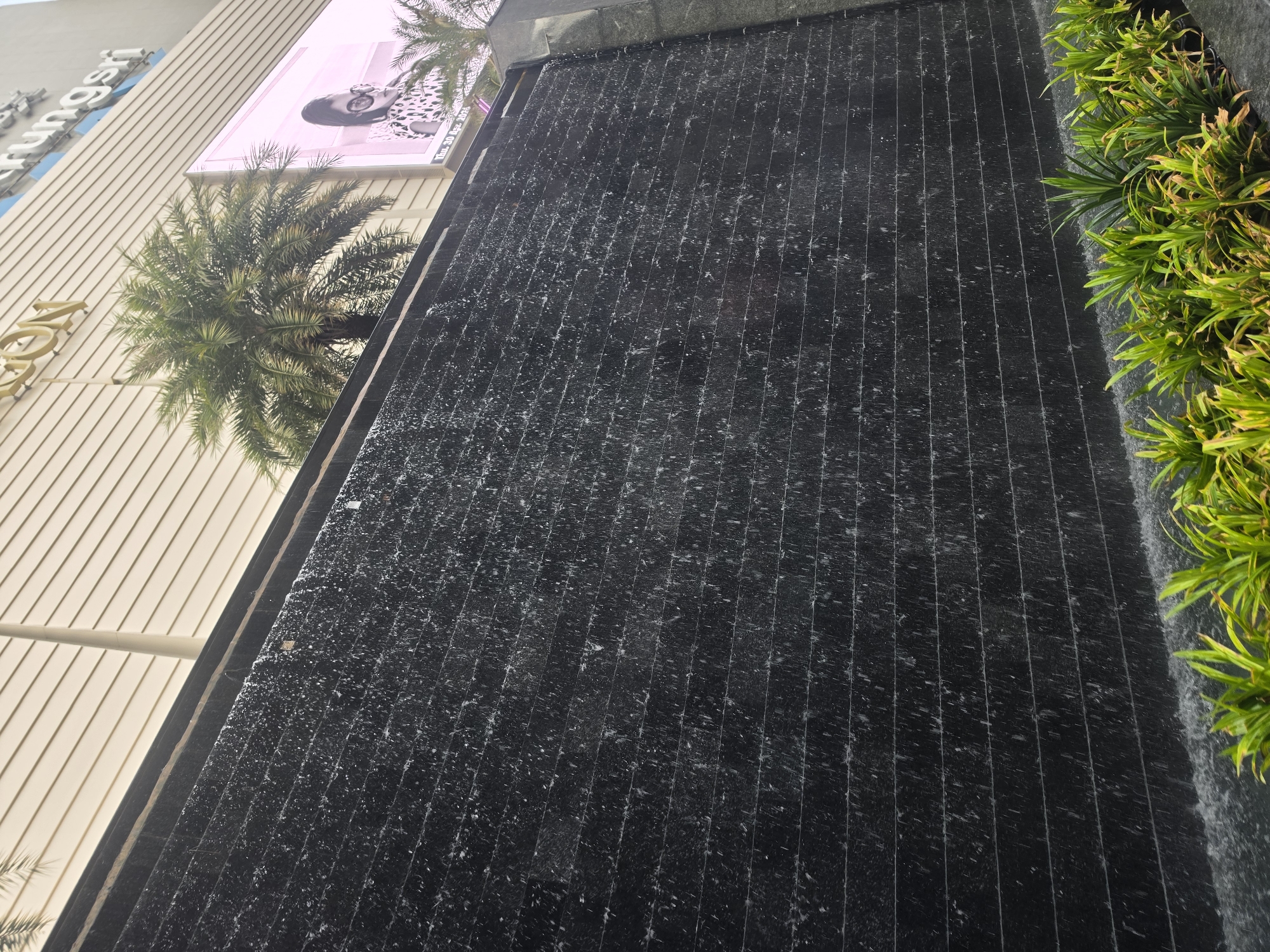 The image shows a modern architectural detail in Bangkok, Thailand. A dark, vertically-ribbed water feature cascades downwards, with water running over its surface. Lush green plants grow along the edge of the water feature. In the background, a section of a building is visible, featuring beige-colored, horizontally-lined cladding. A palm tree and a large advertising billboard displaying a woman's image can also be seen.