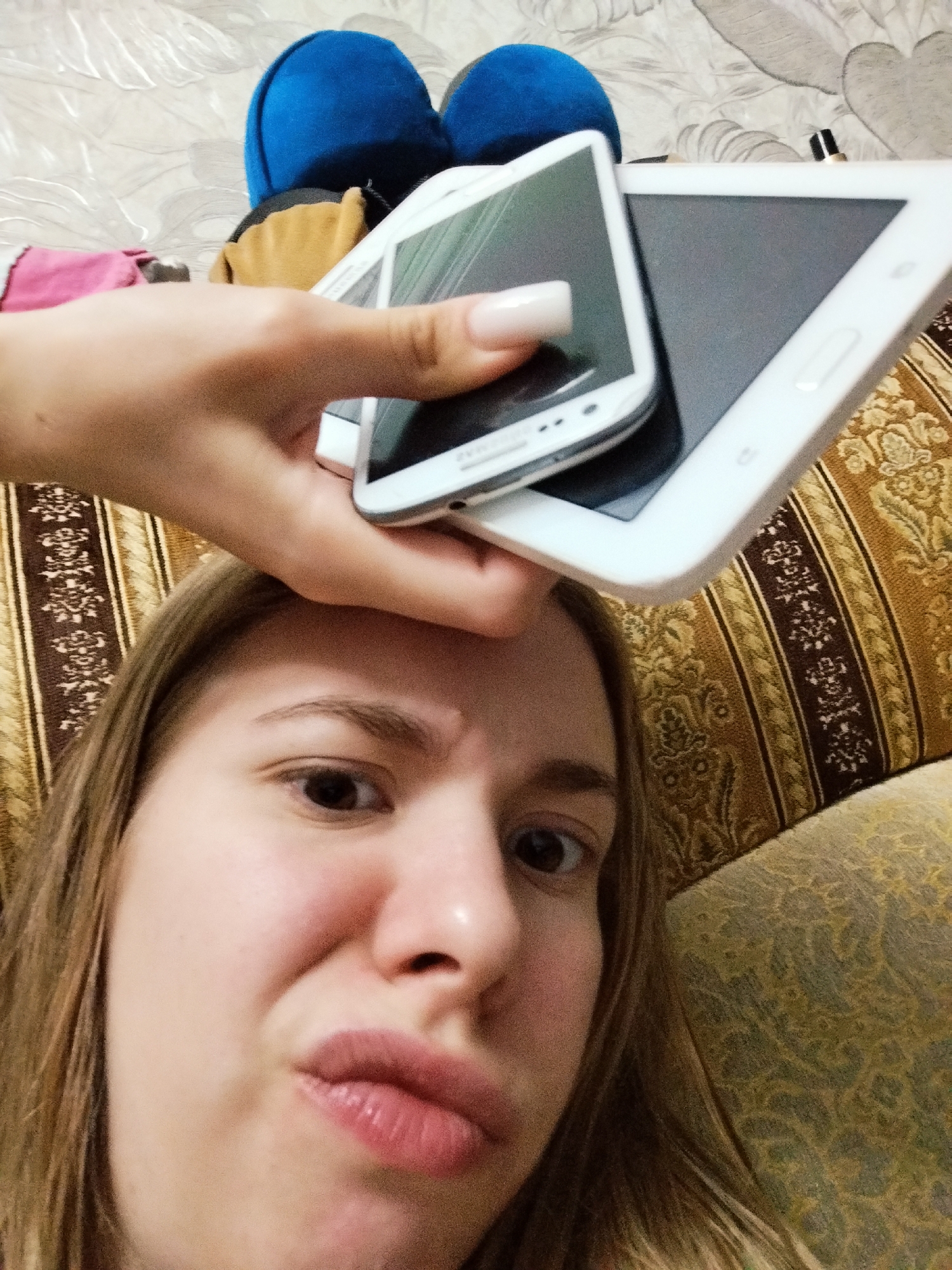 In an indoor setting, a close-up selfie shows a young woman with brown hair. Her mouth is pursed, and her eyes are wide. She is holding two white electronic devices, a smartphone and a tablet, above her face, obscuring part of her forehead. The devices are stacked with the smartphone on top of the tablet. The background includes a textured surface and upholstered furniture with a brown and gold patterned fabric. There are also two blue rounded objects visible in the upper background.