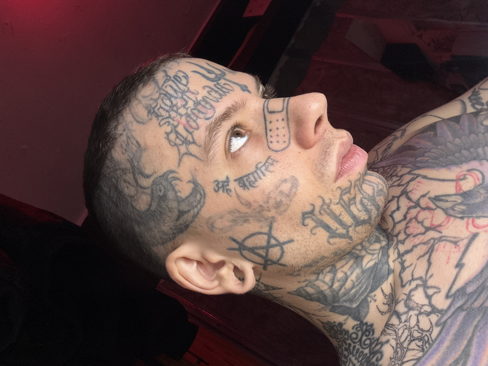 A low-angle, close-up indoor shot depicts a man lying on his back, gazing upwards. He is fair-skinned with a shaved head and numerous tattoos covering his face, neck, and upper chest. The tattoos include various symbols, words in different scripts, and images. He has light-colored eyes. A bandage-like tattoo with small dots is on the side of his nose. The background is out of focus but appears to show parts of a wooden structure or furniture. The lighting is predominantly red. There is no evidence to support the subject is in Buenos Aires, Argentina.