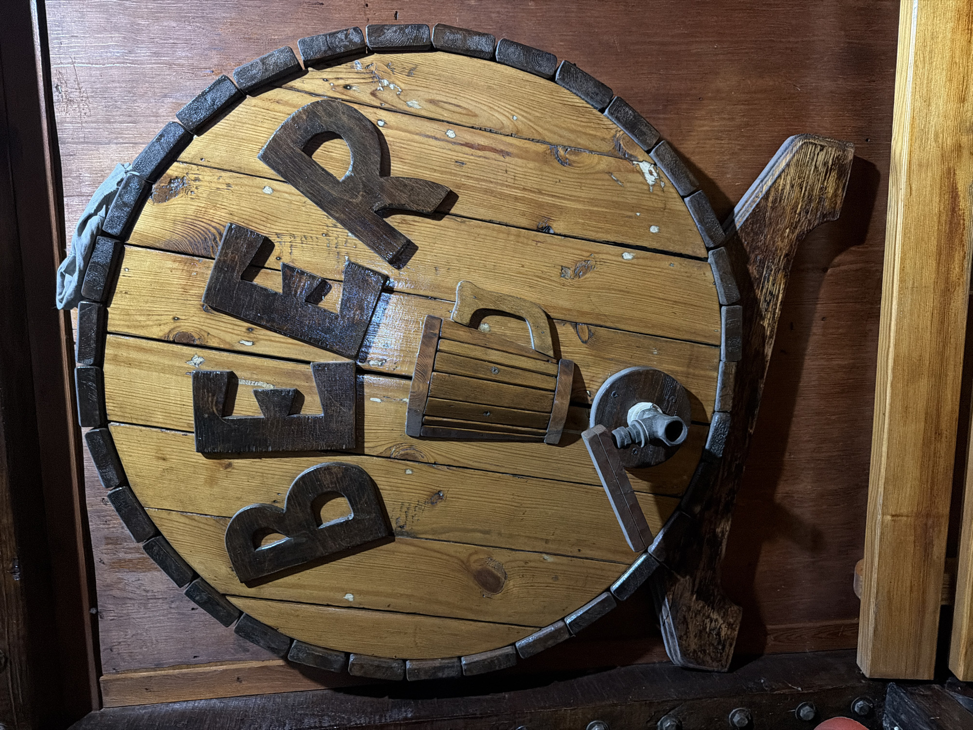 The image shows a wooden barrel-shaped sign with the word "BEER" in large wooden letters. In the center of the barrel, there is a wooden mug with a handle and a spigot. The sign is mounted on a wooden wall, and there is a wooden shelf below it. The lighting is dim, creating shadows. The image is taken from a slightly high angle. There are no people visible in the image. This is a decorative sign that would typically be found in a bar or pub. The location in the city of Semey, Kazakhstan is not identifiable from the image.