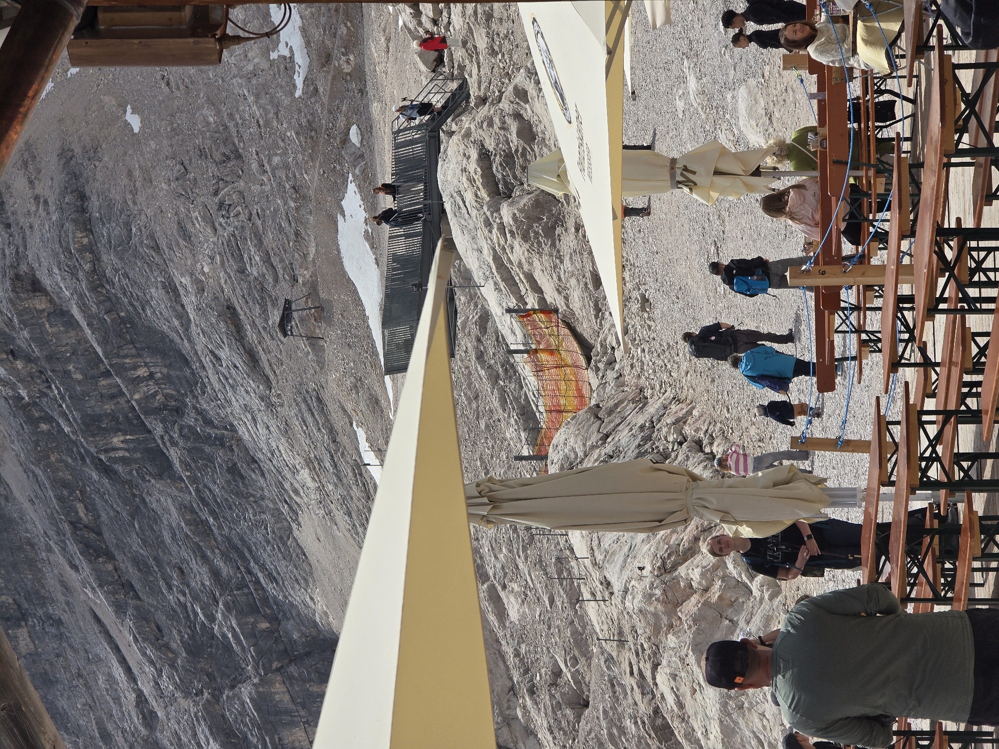 This image depicts a busy outdoor seating area at Zugspitze, Germany's highest peak, located in Garmisch-Partenkirchen. The scene is dominated by a steep, rocky mountain face in the background. In the foreground, there are several picnic tables and benches under large, cream-colored umbrellas, suggesting a restaurant or cafe setting.
Several people are visible throughout the image. A man in a green shirt and a black cap is in the left foreground, facing away from the camera, seemingly observing the surroundings. Behind him, a woman in a black t-shirt with "TEAM" printed on it stands near a folded umbrella. Further back, a group of people are walking on the rocky terrain, including individuals in casual clothing, some with backpacks. One person is wearing a bright blue jacket, and another is in a pink top.
In the middle ground, a raised platform with a metal railing allows people to walk and observe the mountain. Some individuals are seen on this platform and on the stairs leading to it. A bright orange and yellow safety fence is also visible, indicating a protected area.
The environment is characterized by rough, rocky terrain with patches of snow scattered across the mountainside, indicating a high-altitude location. The overall impression is of a tourist destination on a mountain, with visitors enjoying the views and facilities.