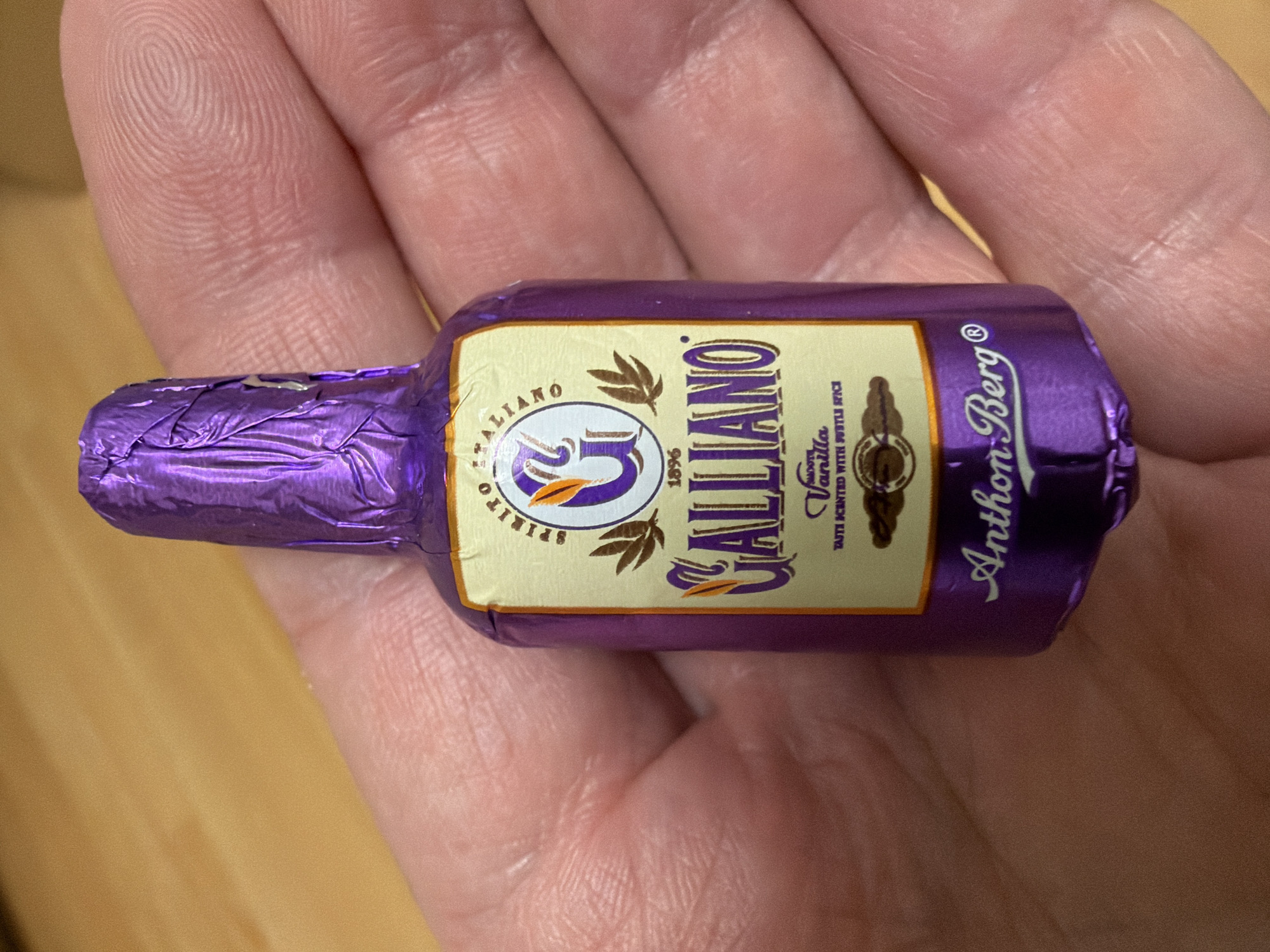 A person is holding a miniature bottle-shaped chocolate wrapped in purple foil. The chocolate is a replica of a Galliano liqueur bottle, with the brand name "GALLIANO" prominently displayed on the label. The label also features the words "SPIRITO ITALIANO," "1896," "Smooth Vanilla," and "TASTE SCENTED WITH SUBTLE SPICE." Below the Galliano branding, the text "Anthon Berg" is visible, indicating the chocolate manufacturer. The chocolate is held in the palm of a hand, with parts of the fingers visible around the object. The background is a plain wooden surface. No specific location within the city of Kyiv, Ukraine, can be determined from the image.