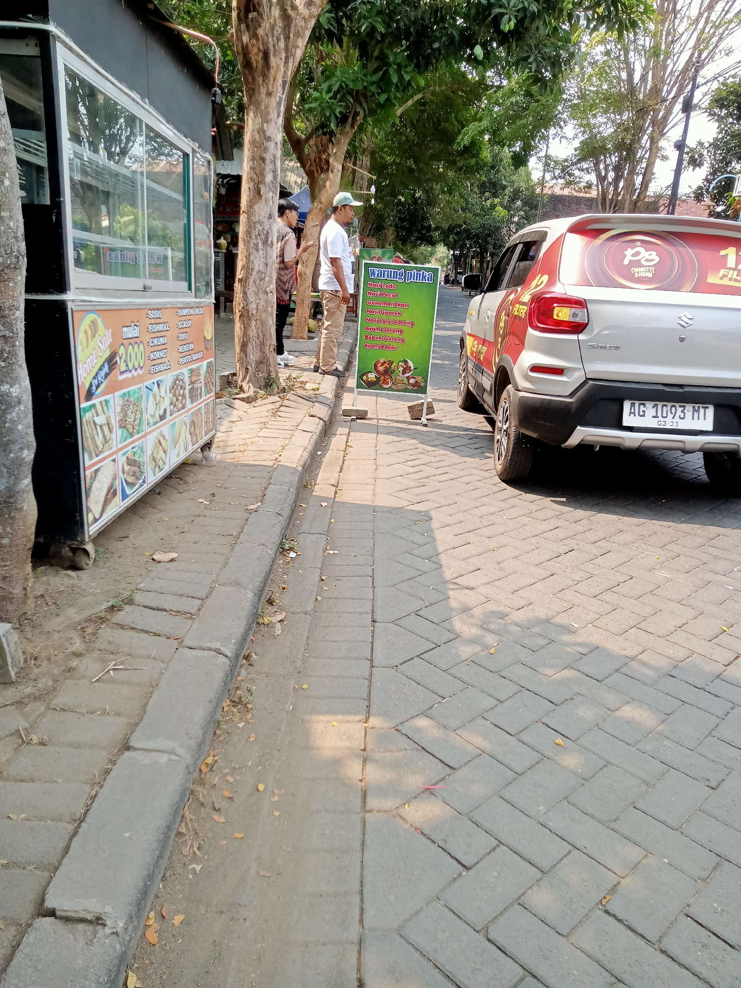 This image depicts a street scene in Tulungagung, Indonesia, likely near a roadside food stall and a parked vehicle. On the left, a food stall with a glass display case is visible. A menu board, which appears to be advertising various food items like "sosis," "fishroll," "dumpling," "nasi pecel," and "ayam goreng," is positioned next to the stall. In front of the stall, there's a price list for "Home Sale" items, with a prominent "2000" indicated.
Two individuals are standing near the stall. One, wearing a light-colored cap and white t-shirt, faces away from the camera. The other person, in a patterned shirt, stands beside them.
To the right of the food stall and people, a silver Suzuki S-presso car is parked. The car has a red and white advertisement or decal on its rear door, featuring the letters "PS" and "12". The license plate reads "AG 1093 MT".
The environment suggests an outdoor setting, with trees providing shade. The ground is paved with interlocking bricks. The lighting indicates it is daytime, with shadows cast by the trees and objects. The overall impression is a typical street scene with local businesses and everyday activities in Tulungagung.