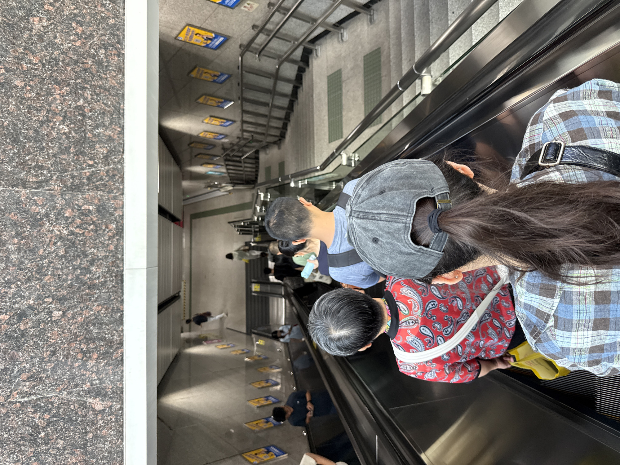 In Tianhe District, China, an overhead view captures people on an escalator descending into a subway station. The escalator is flanked by stairs with metal railings. The walls of the station are adorned with yellow posters featuring portraits and Chinese text. In the foreground, a woman with a gray baseball cap and dark hair pulled into a ponytail is visible from behind, wearing a blue and white plaid shirt. Ahead of her, an elderly woman with gray hair is on the same escalator, wearing a red shirt with a paisley pattern and carrying a yellow bag. Other individuals are seen further down the escalator and on the stairs, most dressed in casual attire. The lighting in the station appears to be artificial.